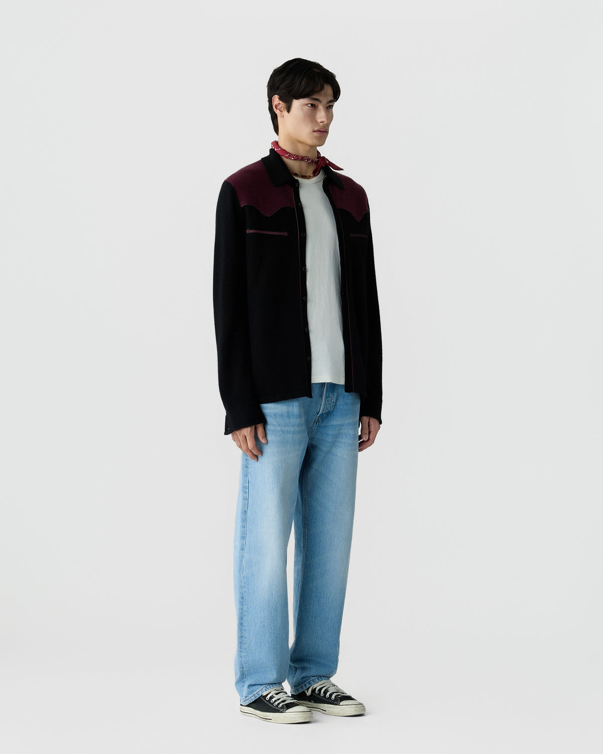 A young man stands against a plain white background wearing a black and burgundy long-sleeve shirt over a white t-shirt, light blue jeans, a red neck scarf, and black sneakers.