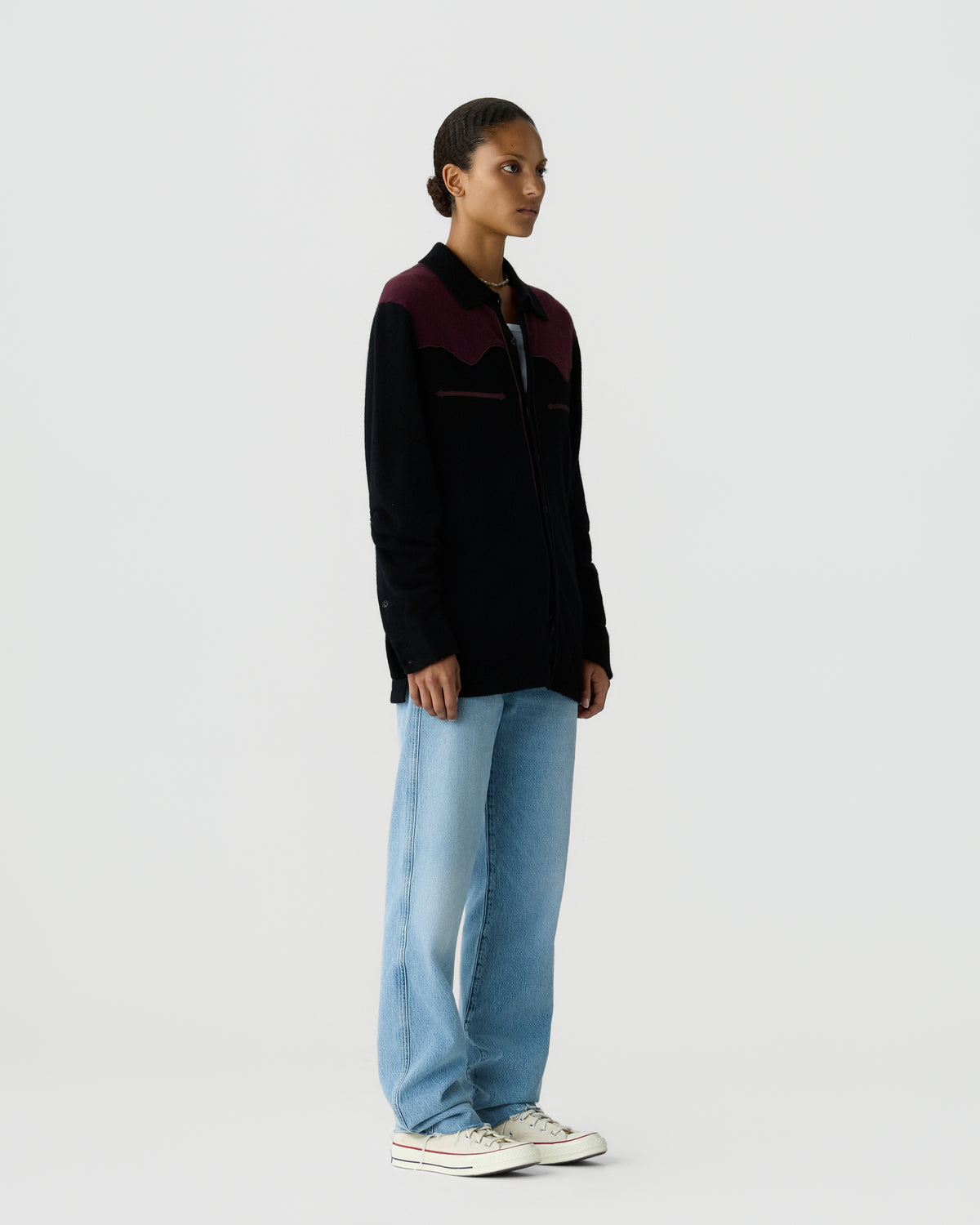 Side view of a person wearing a black and dark purple sweater with light blue jeans and white sneakers standing against a plain light background.