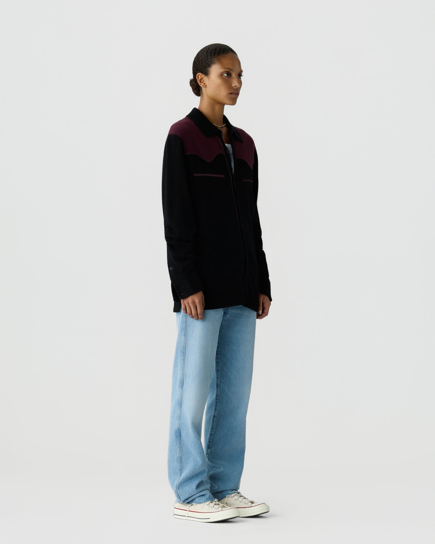 Side view of a person wearing a black and dark purple sweater with light blue jeans and white sneakers standing against a plain light background.