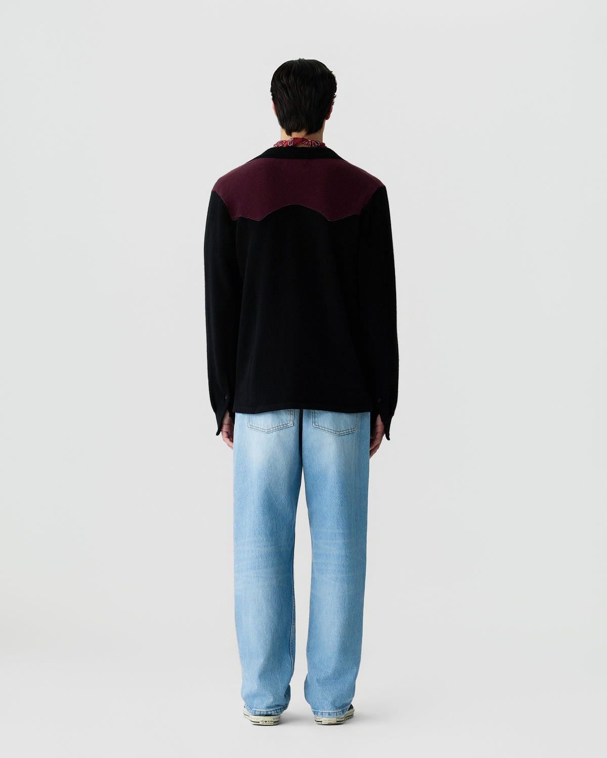 Man standing facing away wearing a black and dark purple sweater with light blue jeans against a plain white background.