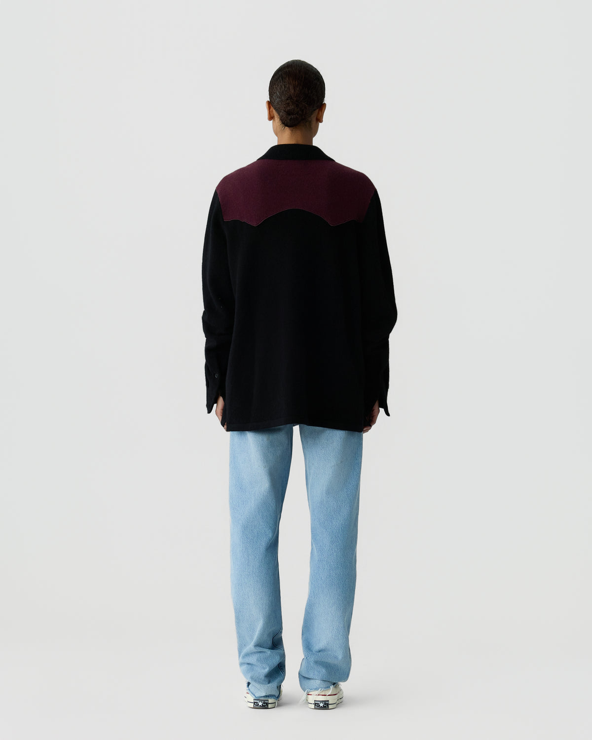 Person wearing a black and dark purple jacket with a wavy color transition on the back, paired with light blue jeans and sneakers, standing against a plain light background.