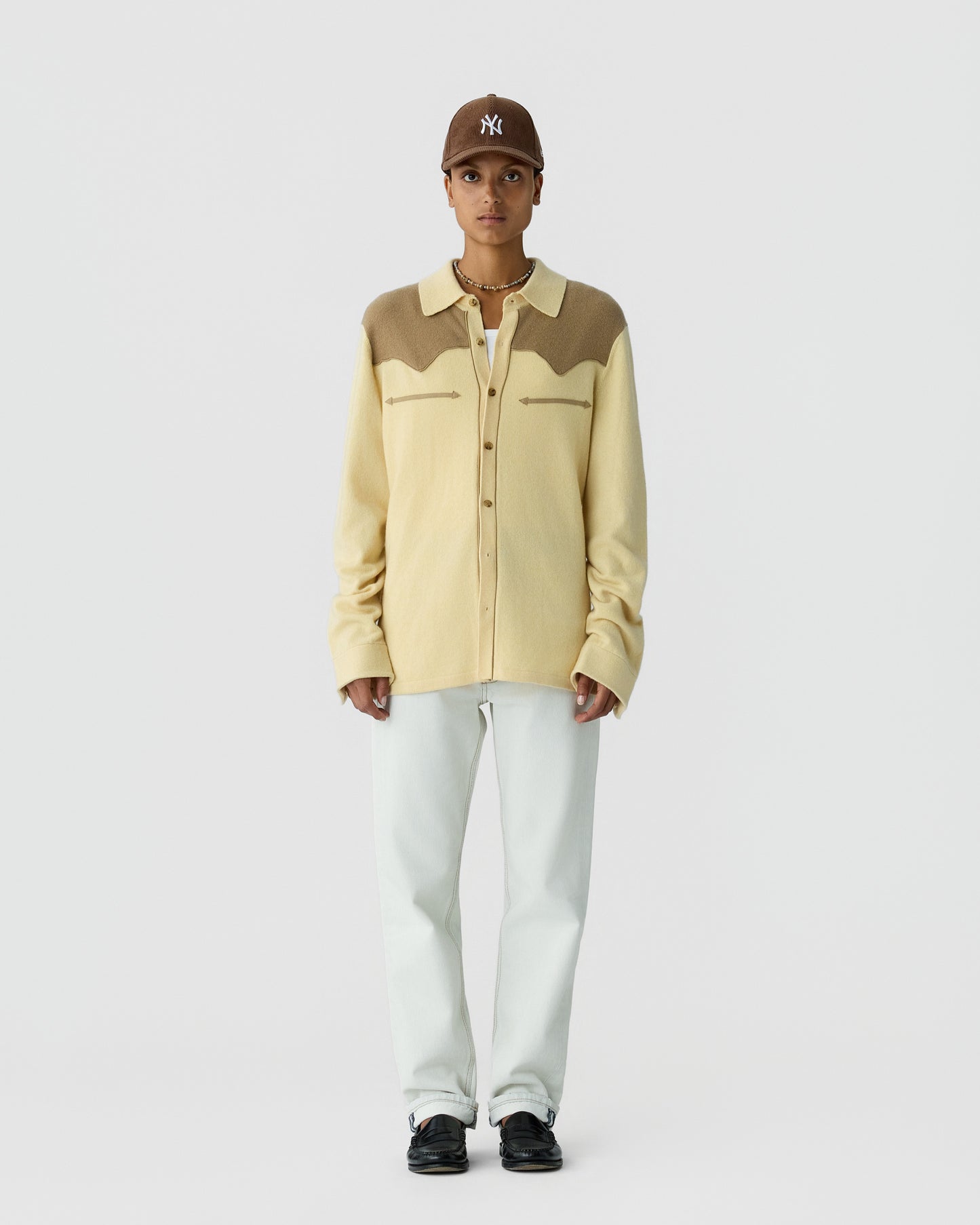 Person wearing a color-block cashmere shirt in cream and brown, a brown baseball cap, white pants, and black shoes, standing against a plain white background.