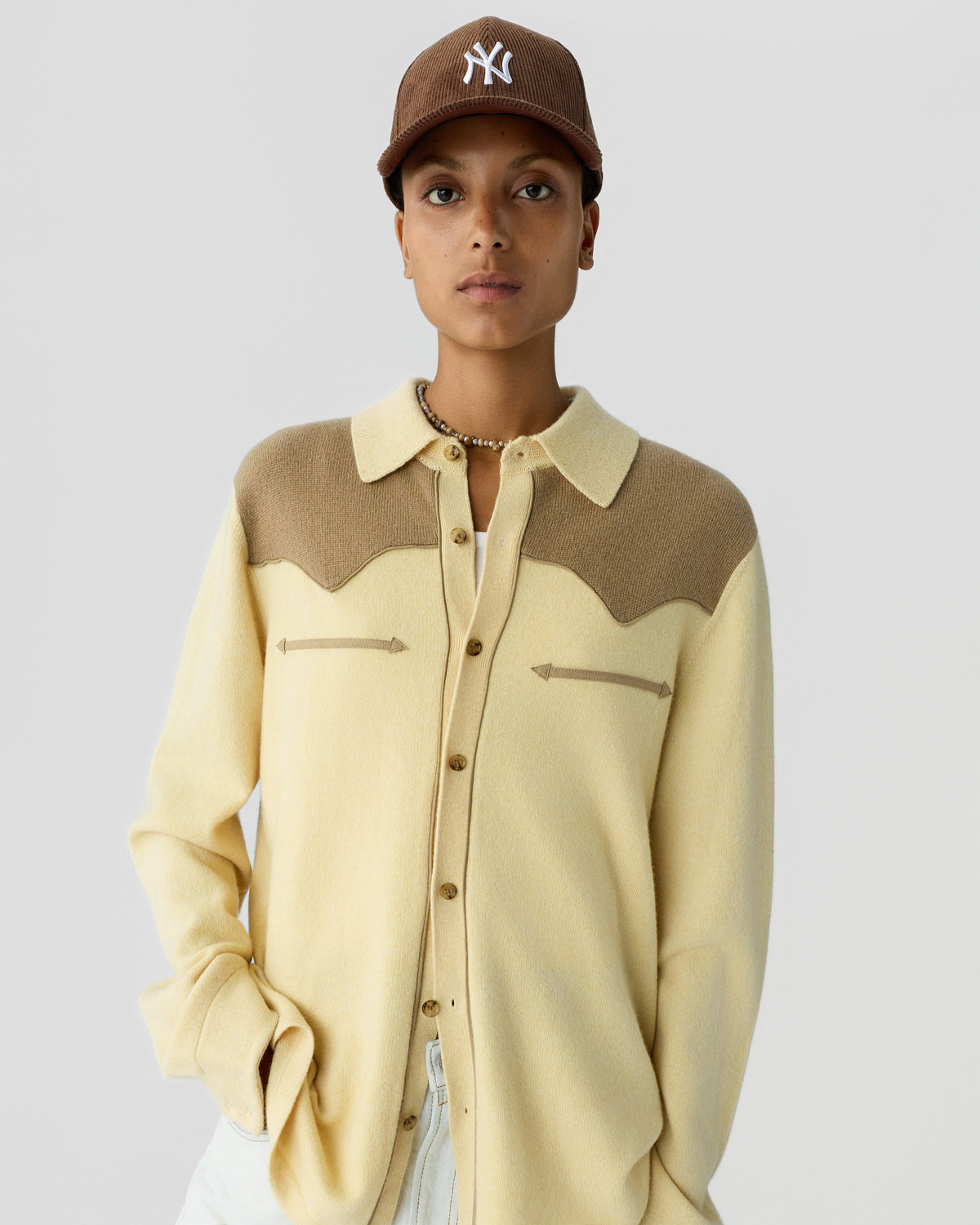 Person wearing a beige and brown long-sleeve button-up shirt with a collar and arrow-shaped details on the chest, paired with a brown baseball cap.