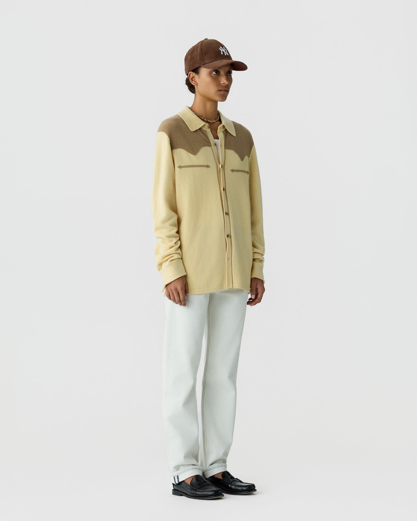 Person wearing a beige and brown long-sleeve buttoned cardigan with a collar, white pants, black shoes, and a brown baseball cap standing against a plain light background.