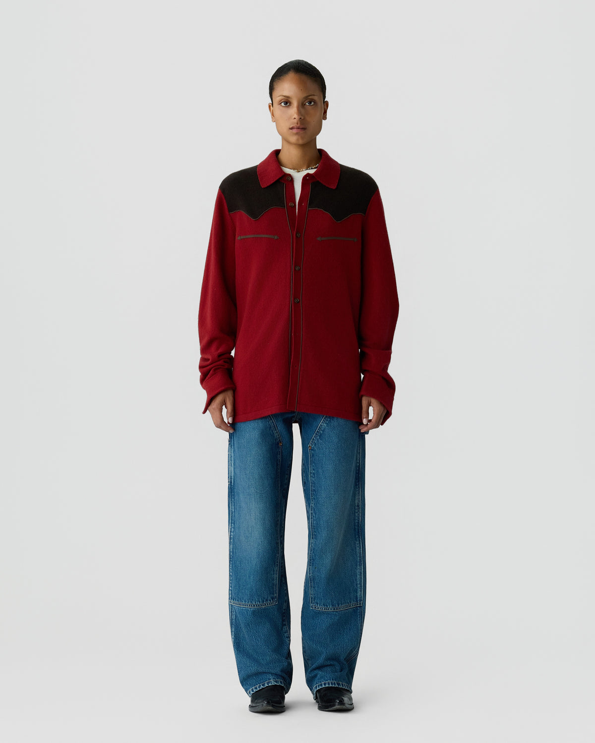 Person wearing a red and black long-sleeve button-up shirt with denim jeans and black shoes, standing against a plain light gray background.