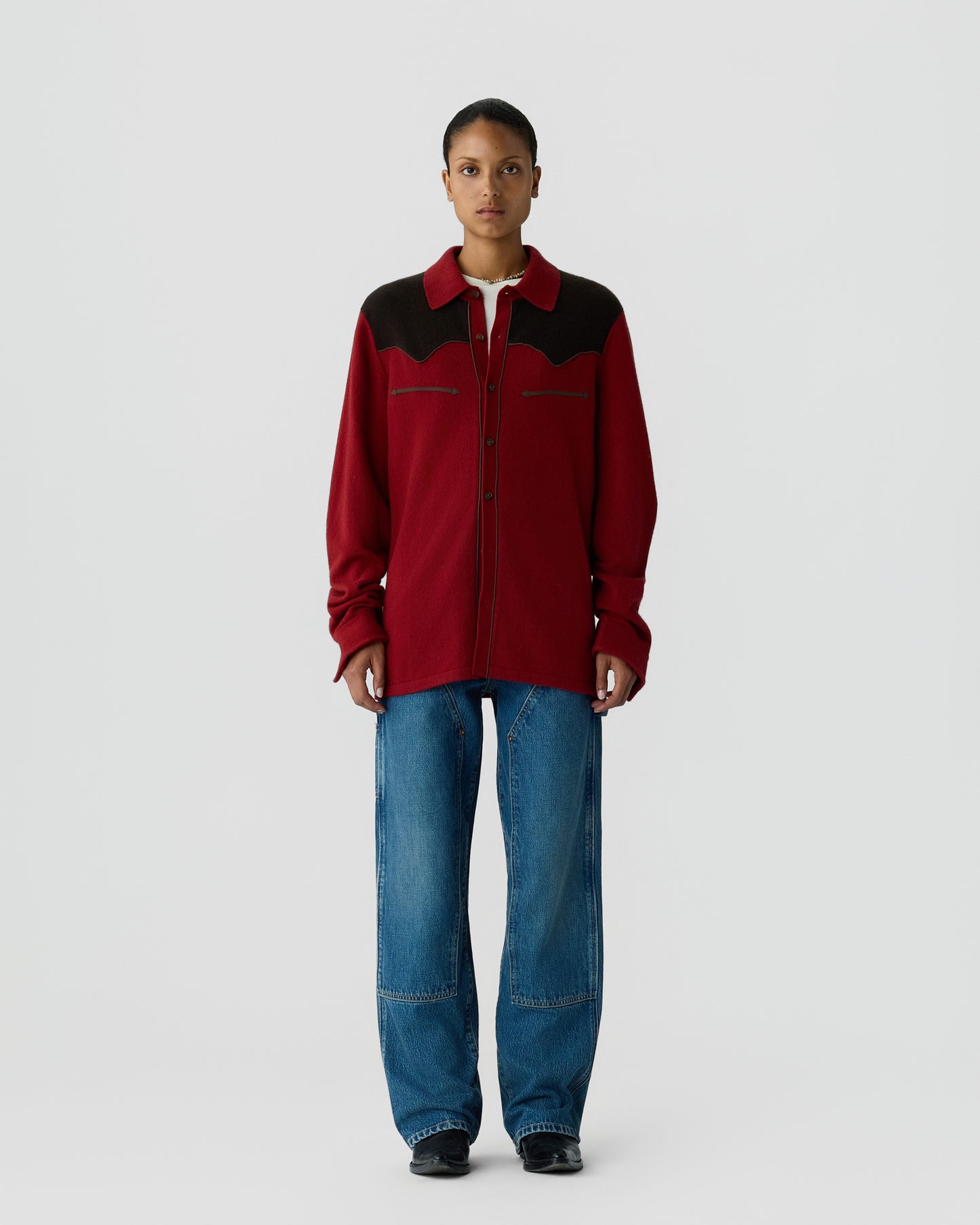 Person wearing a red and black long-sleeve button-up shirt with denim jeans and black shoes, standing against a plain light gray background.