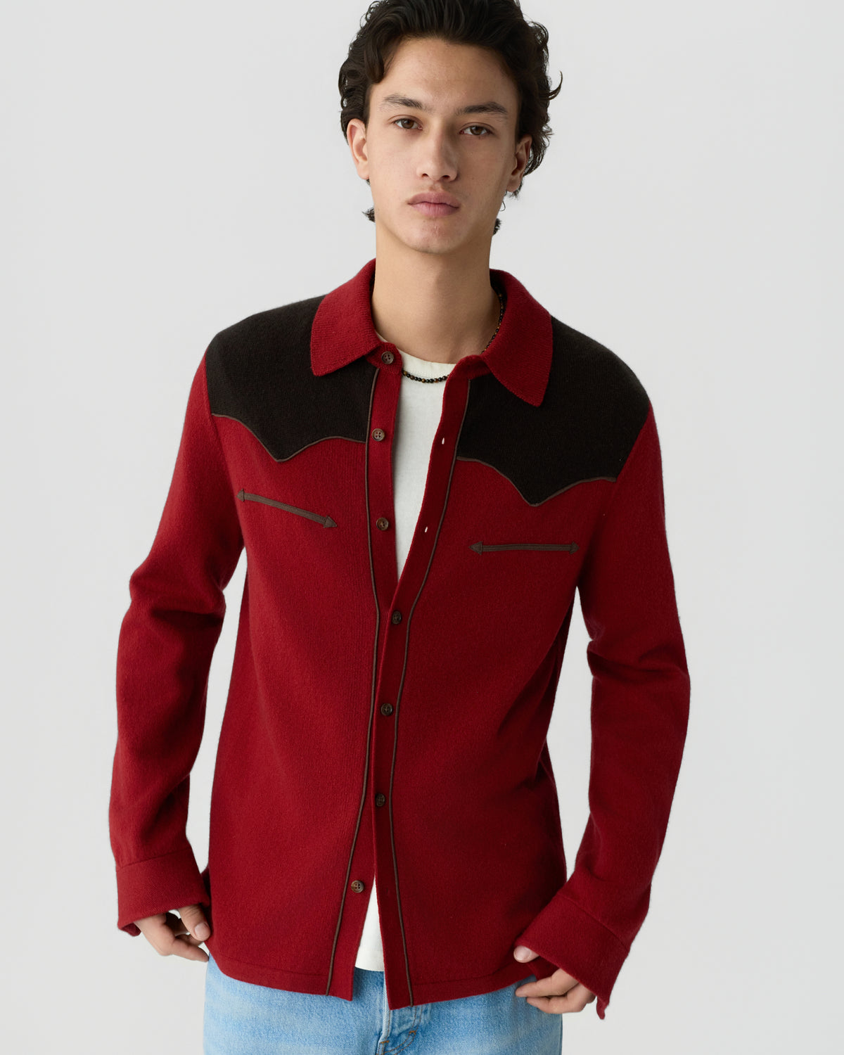 Young man wearing a red and black button-up wool shirt jacket over a white t-shirt and light blue jeans against a plain gray background.