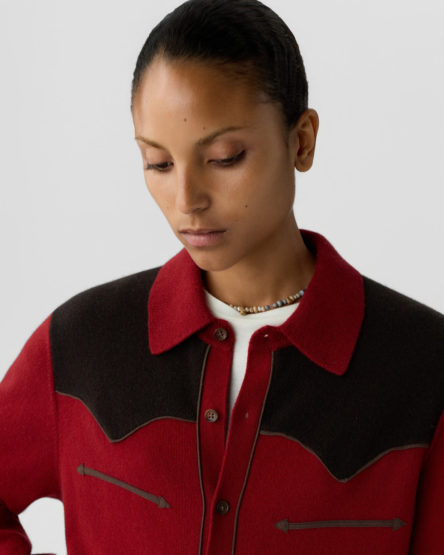 Woman with tied-back hair wearing a red and dark brown wool jacket with button closure and a beaded necklace, looking downward against a plain gray background.