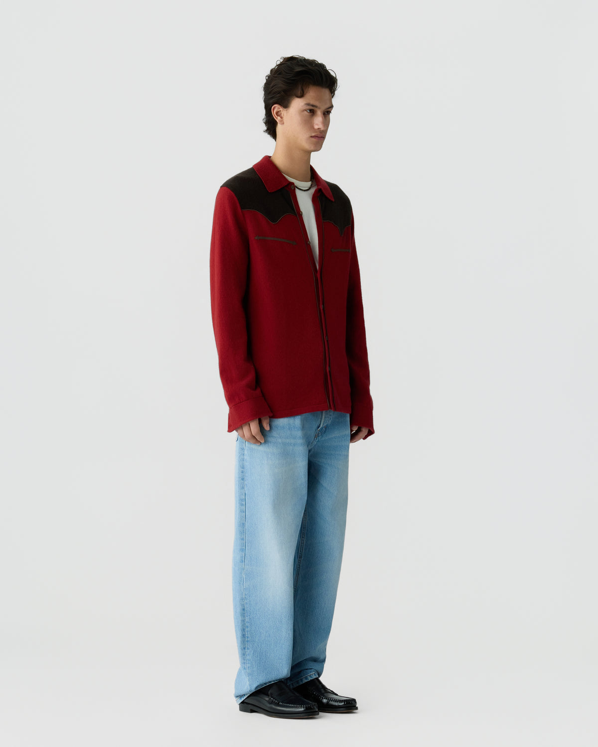 Young man standing wearing a red and dark brown jacket over a white shirt, light blue wide-leg jeans, and black shoes against a plain light background.