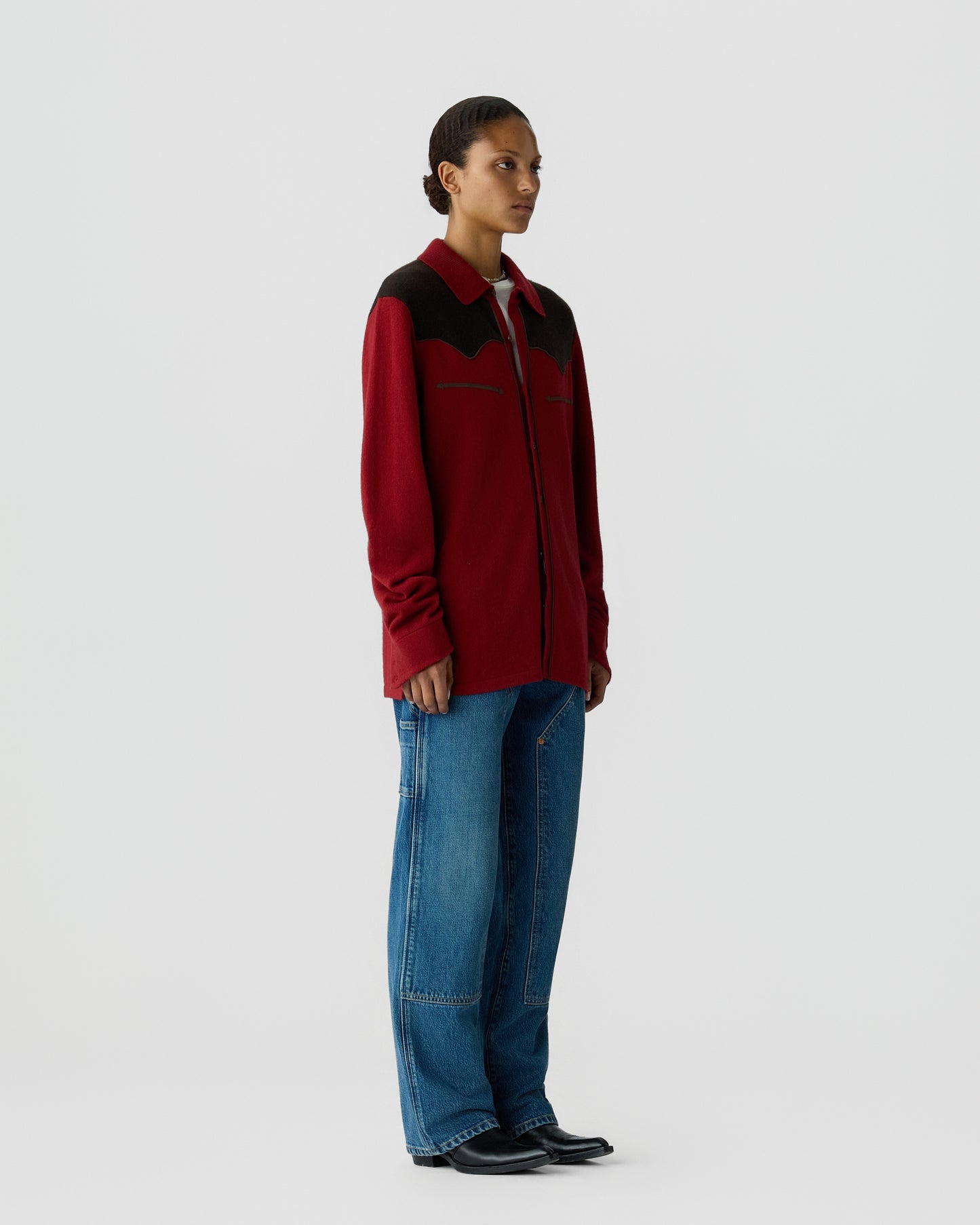 Side view of a person wearing a long-sleeved red and black shirt with a collar, blue wide-leg jeans, and black shoes, standing against a plain light background.
