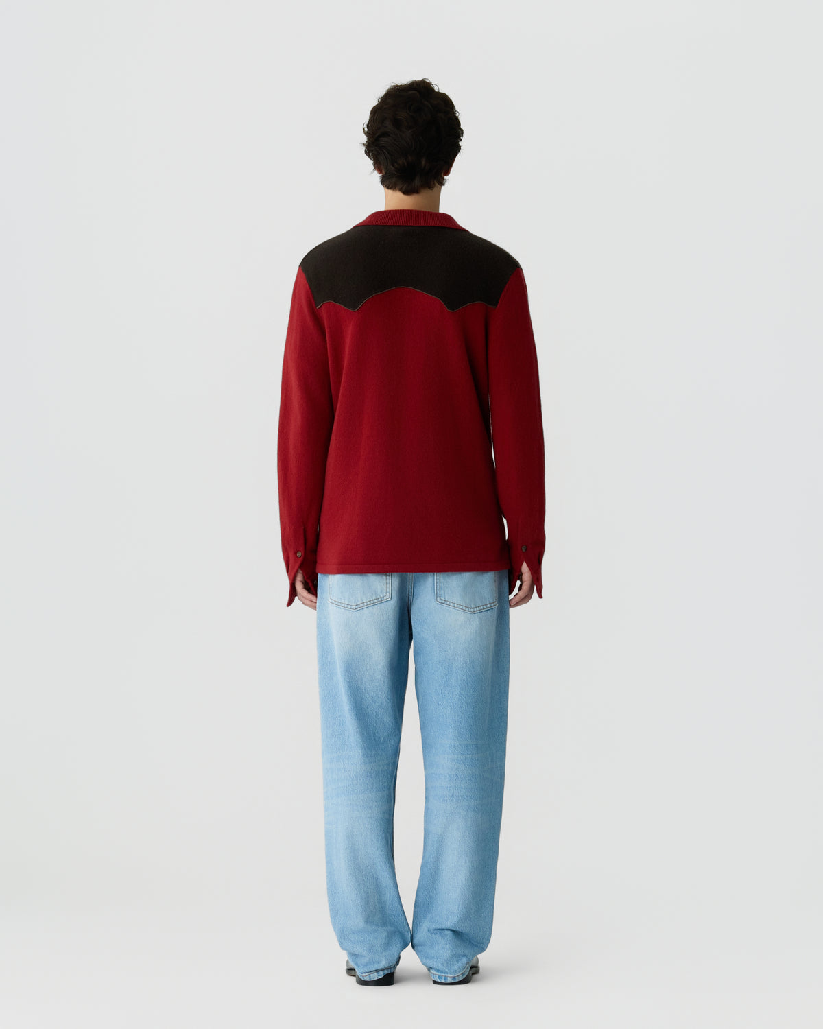 Man viewed from behind wearing a long-sleeve red and black shirt with a scalloped design across the shoulders, paired with light blue jeans against a plain white background.