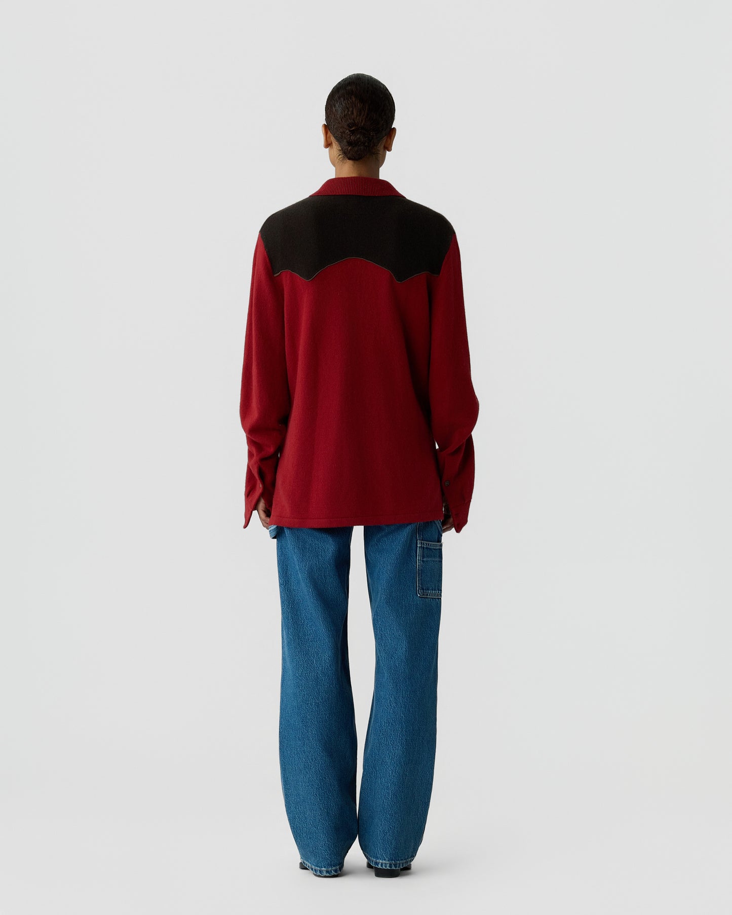 Person standing facing away wearing a long-sleeve burgundy and black top paired with loose blue jeans against a plain light gray background.
