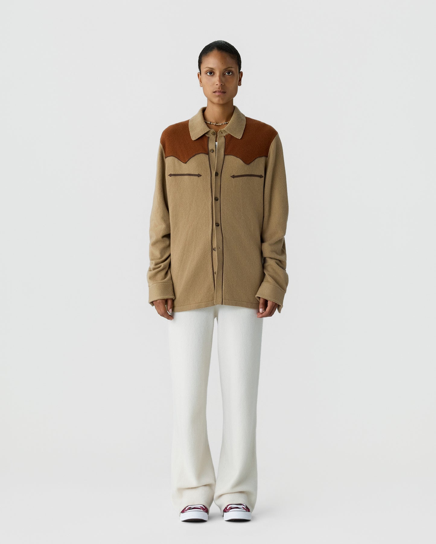 A woman stands against a plain white background wearing a beige and brown button-up shirt with two chest pockets, white wide-leg pants, and white sneakers with red accents.