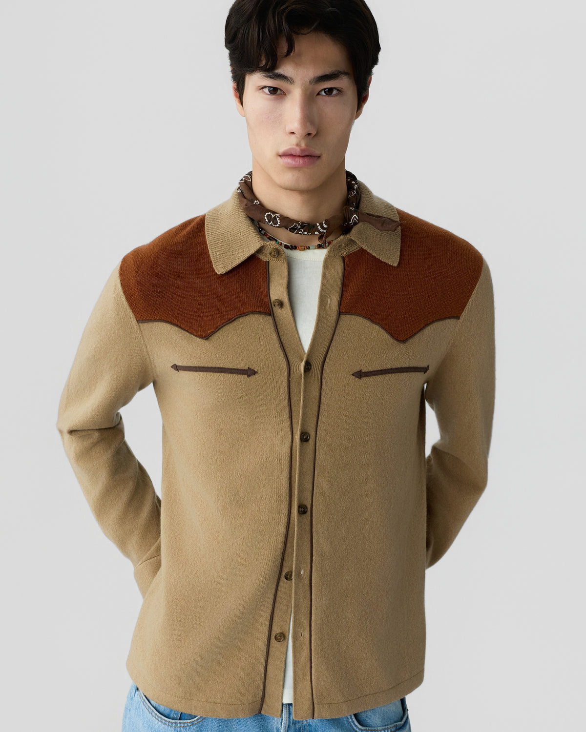 Young man wearing a beige and brown button-up wool shirt with chest pocket details and a patterned neck scarf against a plain gray background.