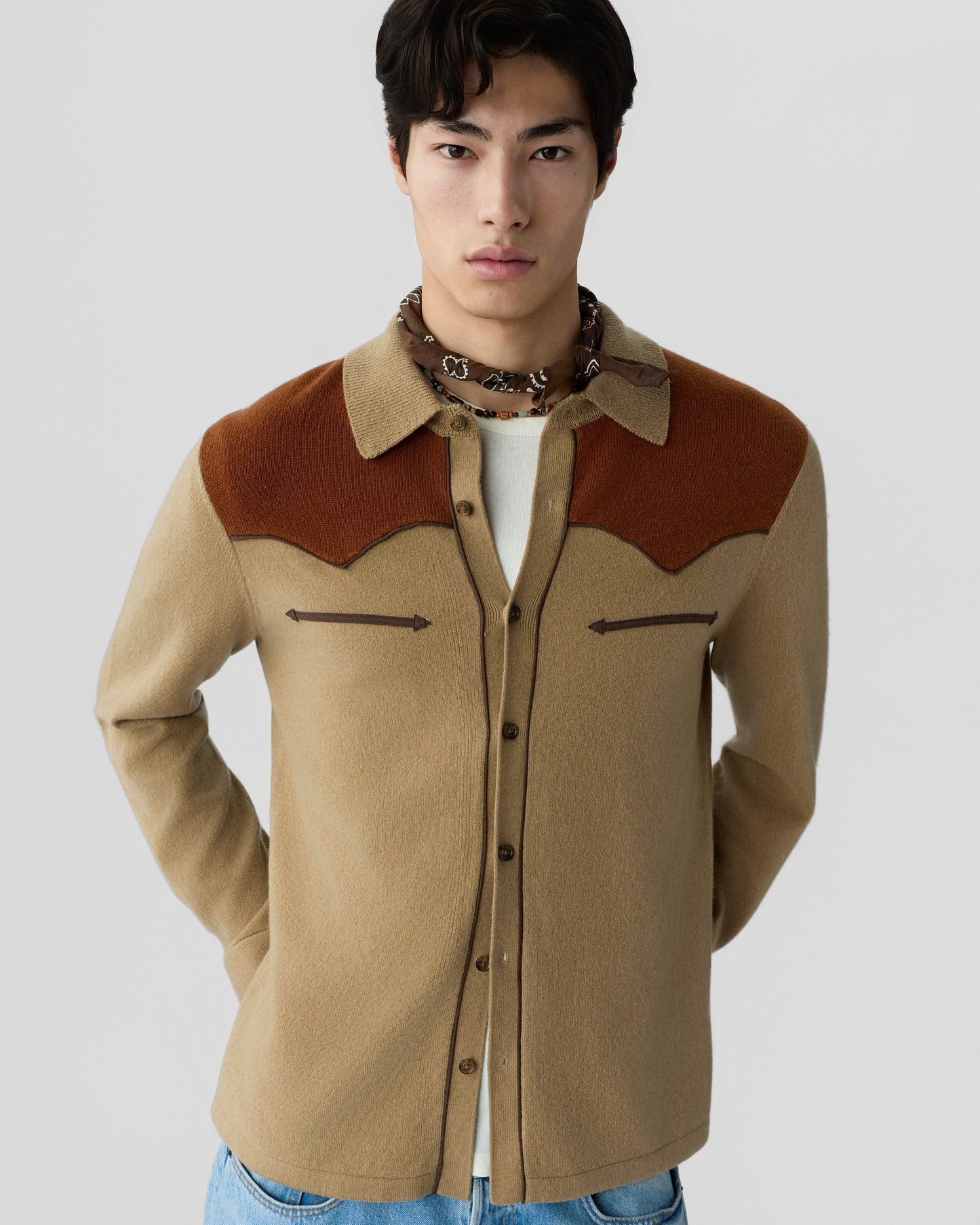 Young man wearing a beige and brown button-up wool shirt with chest pocket details and a patterned neck scarf against a plain gray background.