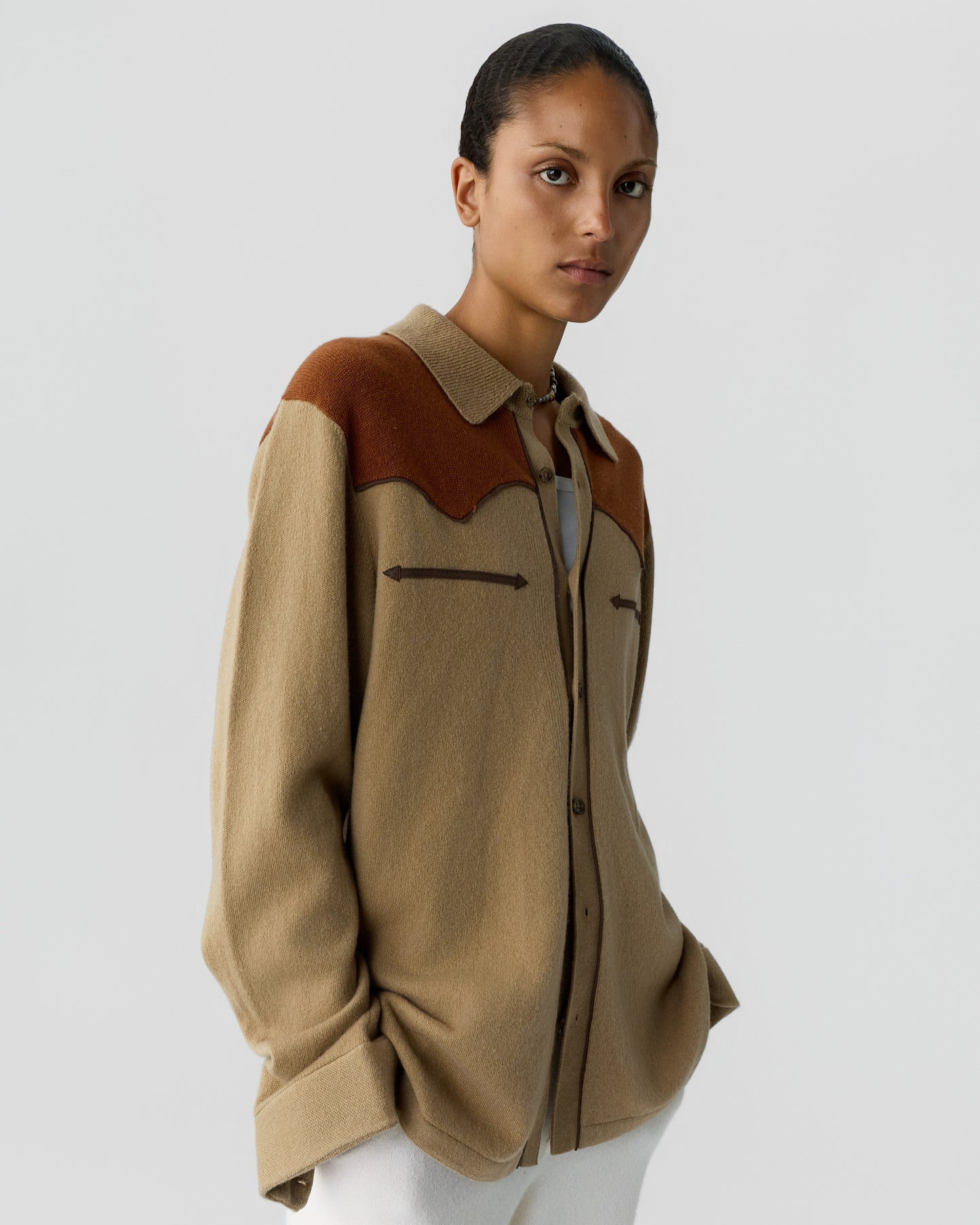 Woman wearing a loose-fitting beige and brown long-sleeve shirt with a collar and arrow-shaped stitching detail on the chest, standing against a plain light gray background.