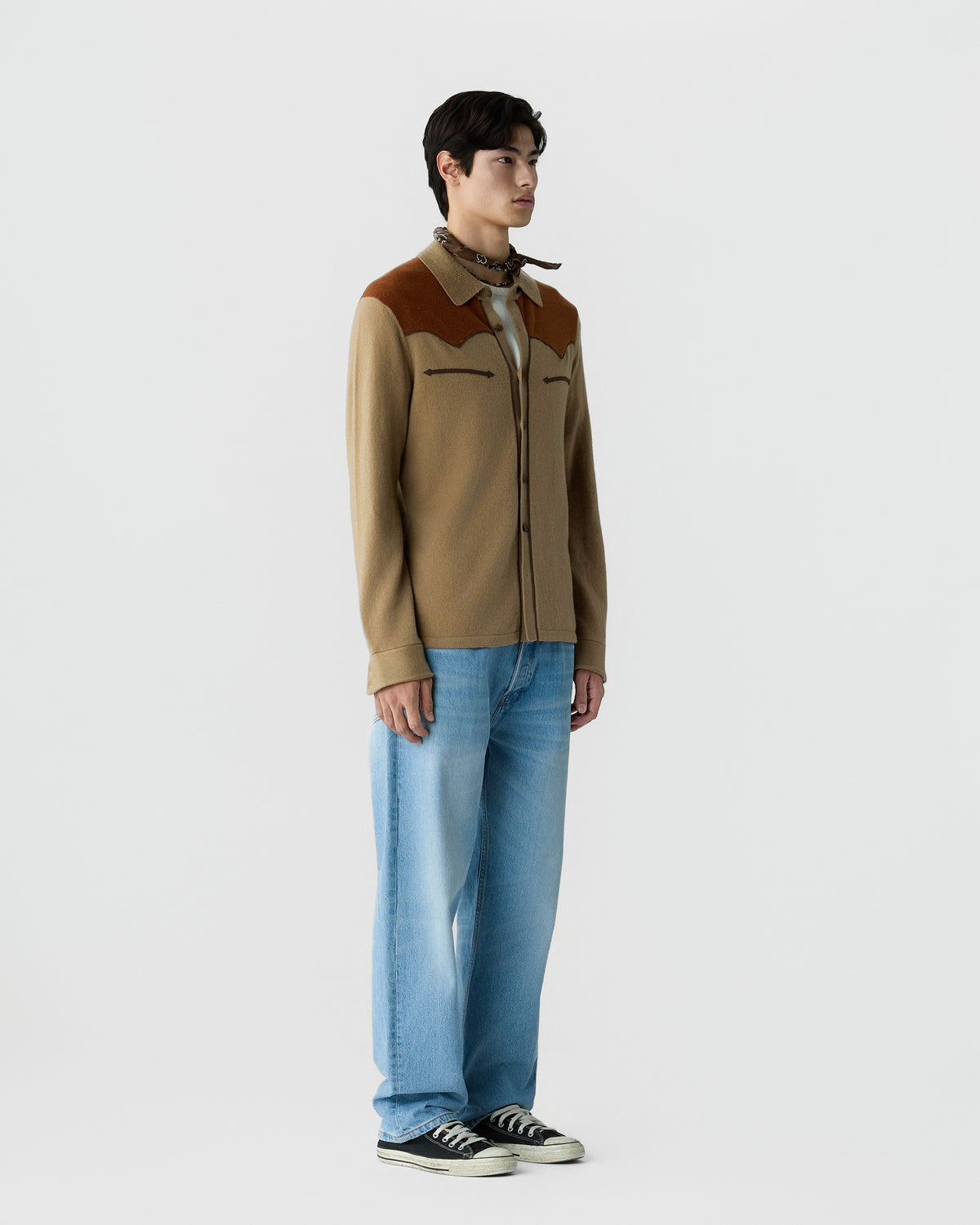 A young man stands against a plain white background wearing a beige and brown jacket with western-style yoke details, light blue wide-leg jeans, black low-top sneakers, and a patterned neck scarf.