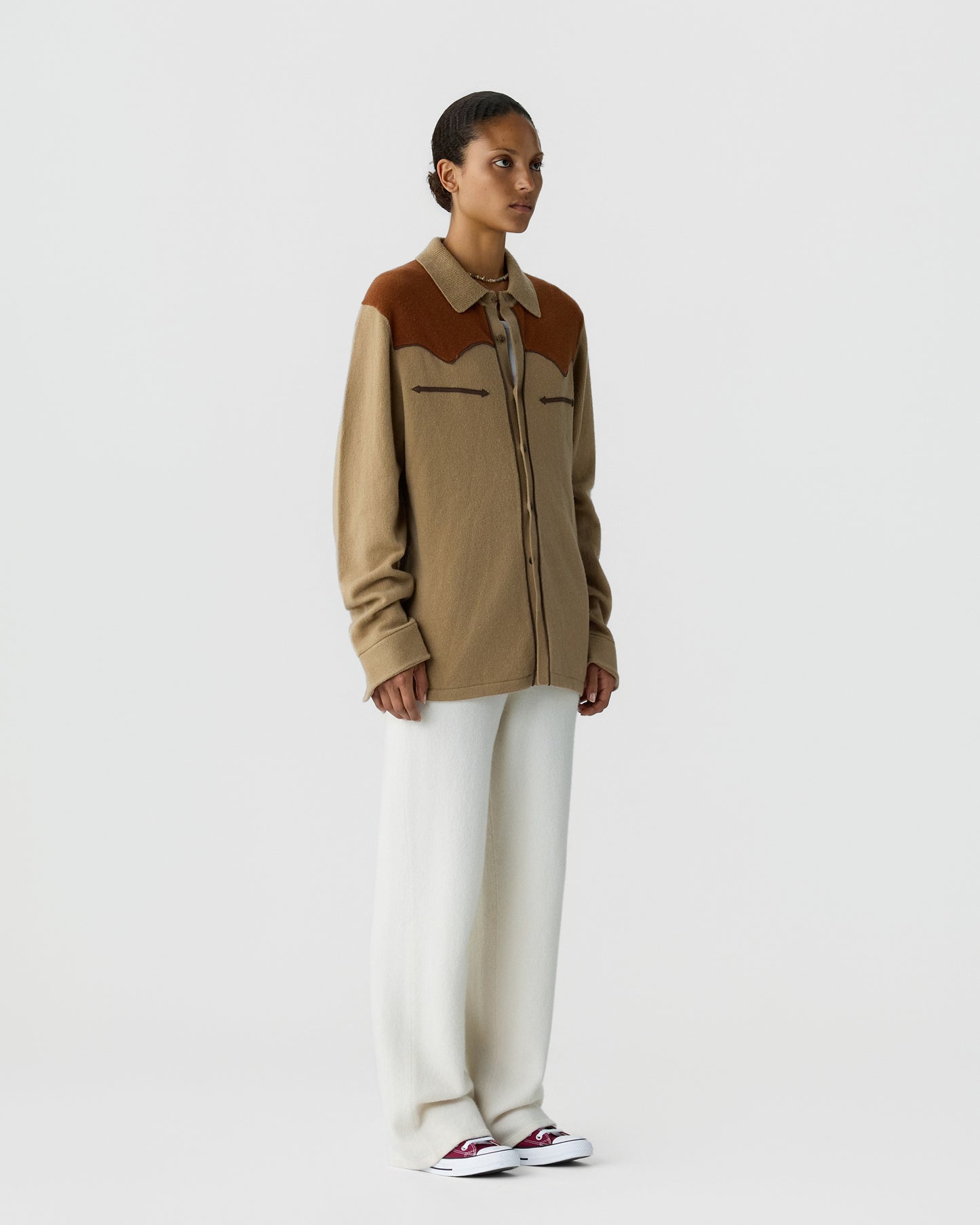 Woman wearing a beige and brown long-sleeve button-up shirt paired with wide-leg white pants and white sneakers, standing against a plain white background.