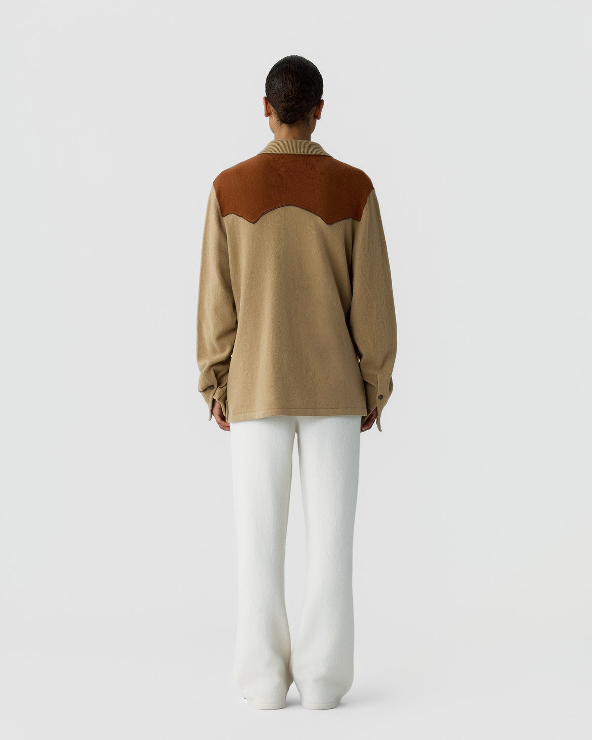 Person seen from the back wearing a beige long-sleeve shirt with a rust-colored yoke detail and white loose-fitting pants against a plain light gray background.