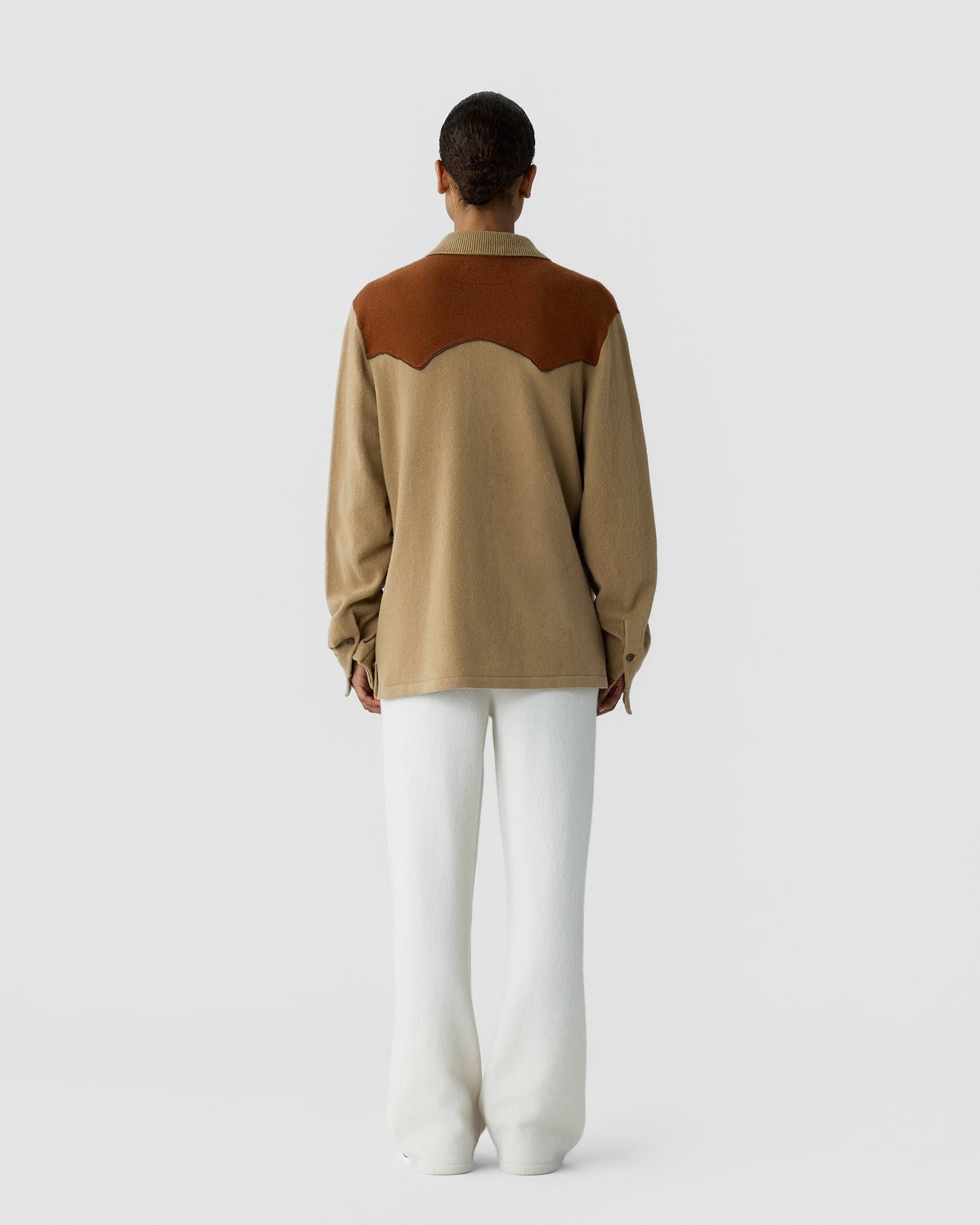 Person seen from the back wearing a beige long-sleeve shirt with a rust-colored yoke detail and white loose-fitting pants against a plain light gray background.