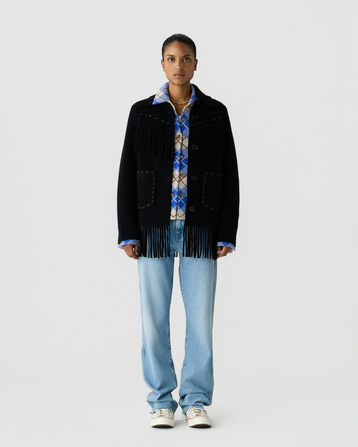 Full length portrait of a person wearing a black fringed jacket over a blue patterned shirt with light blue jeans and white sneakers against a pale gray background