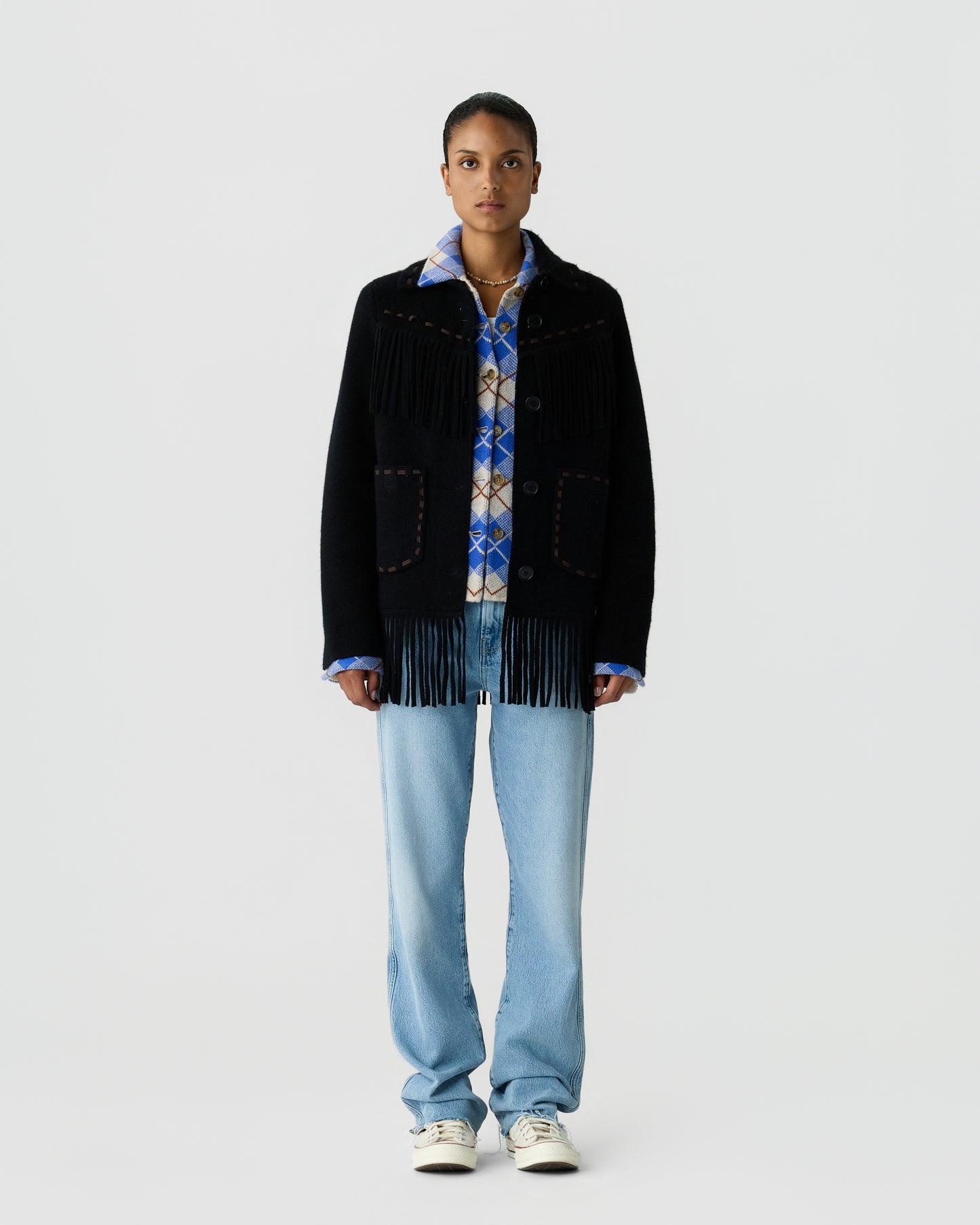 Full length portrait of a person wearing a black fringed jacket over a blue patterned shirt with light blue jeans and white sneakers against a pale gray background