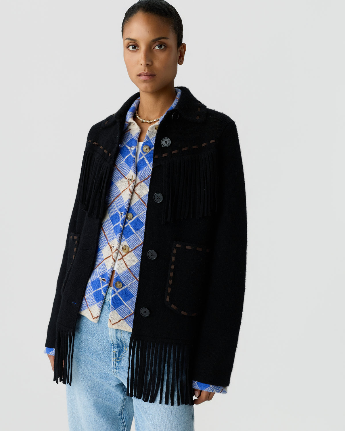 Woman wearing a black fringed jacket over a blue and white argyle sweater with light blue jeans against a plain gray background