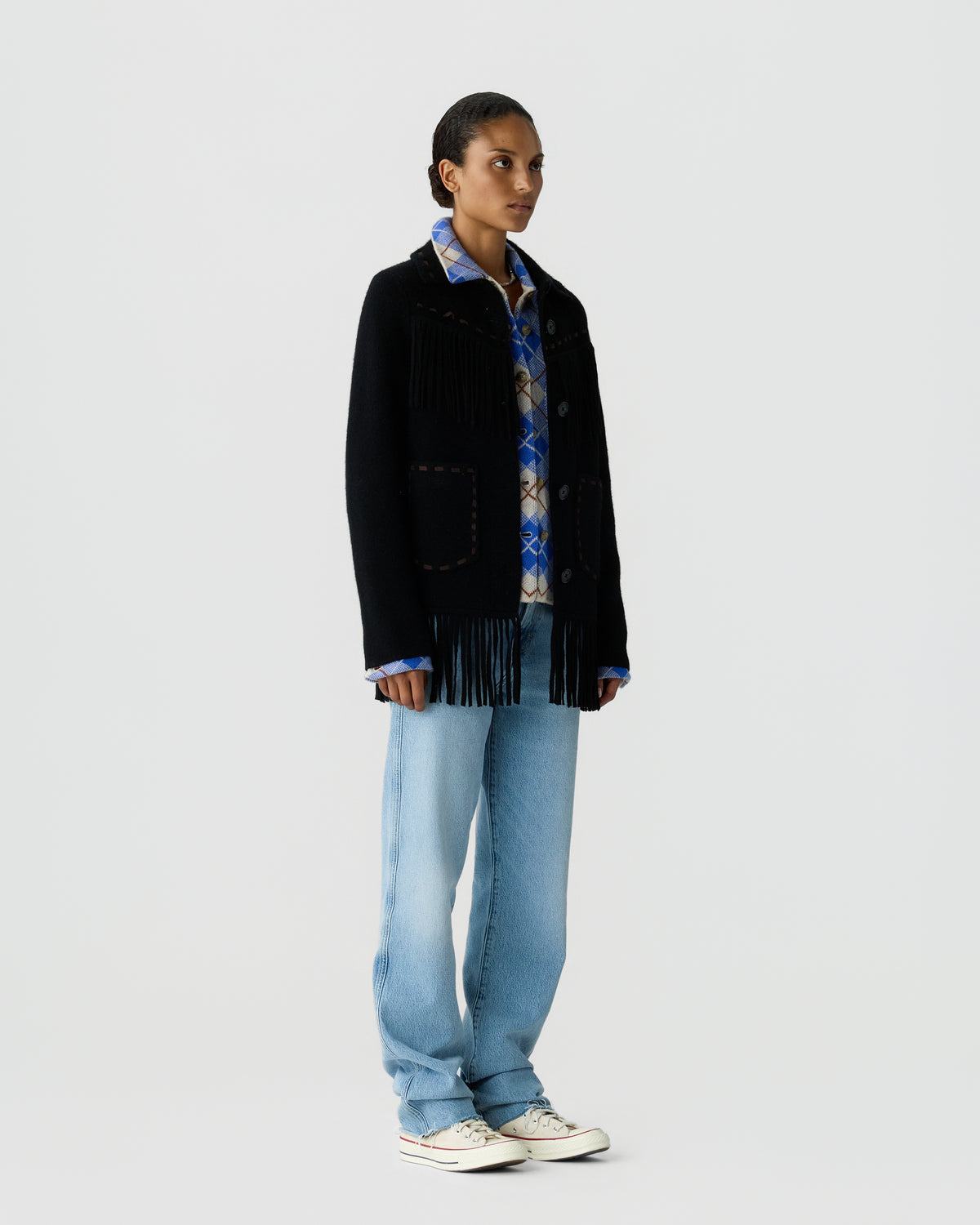 Full length view of a model wearing a black fringed jacket over a patterned blue shirt with loose light wash jeans and white sneakers on a pale gray background