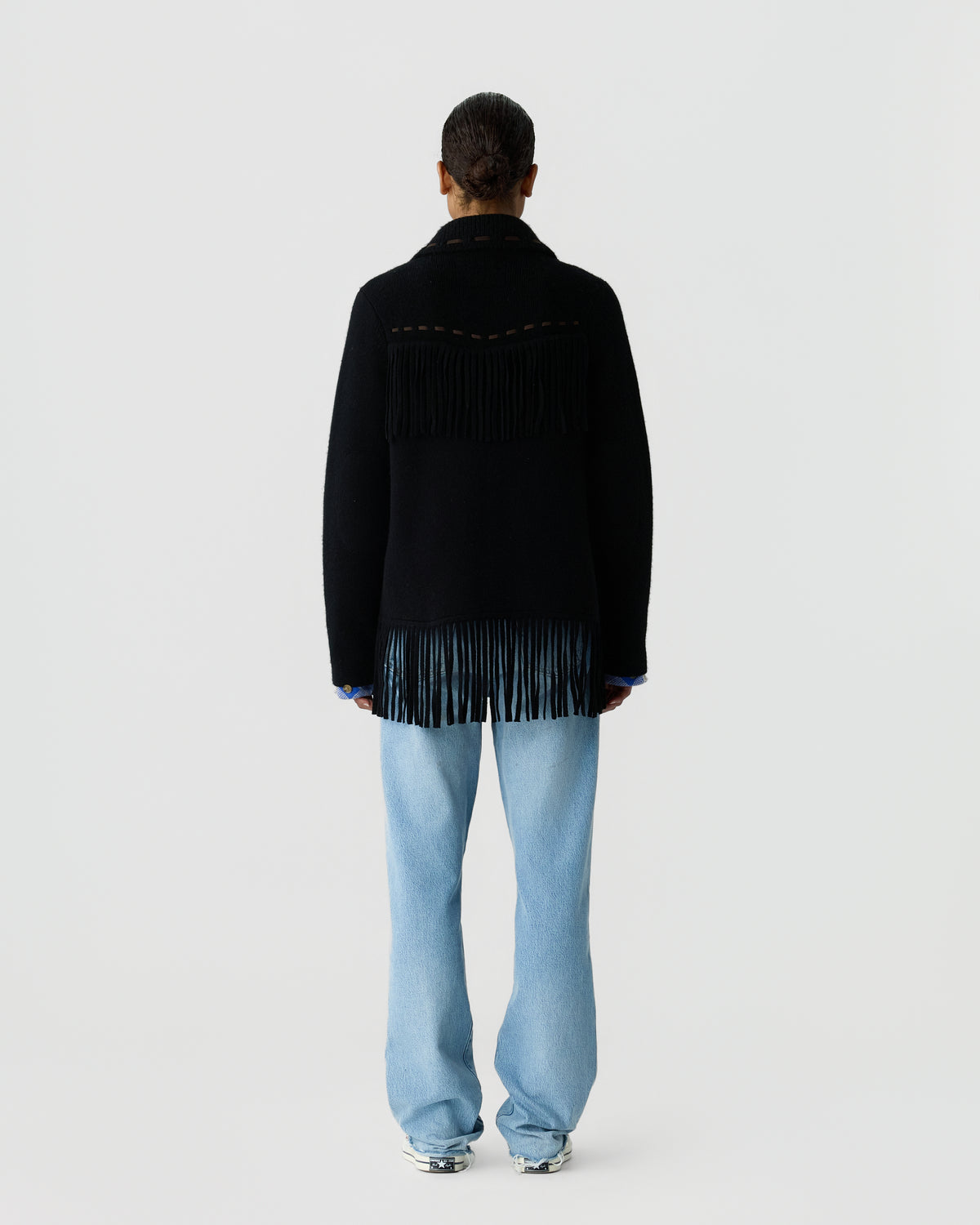 Back view of a person wearing a black fringed sweater with a high collar, light blue wide-leg jeans, and silver shoes against a plain light gray background