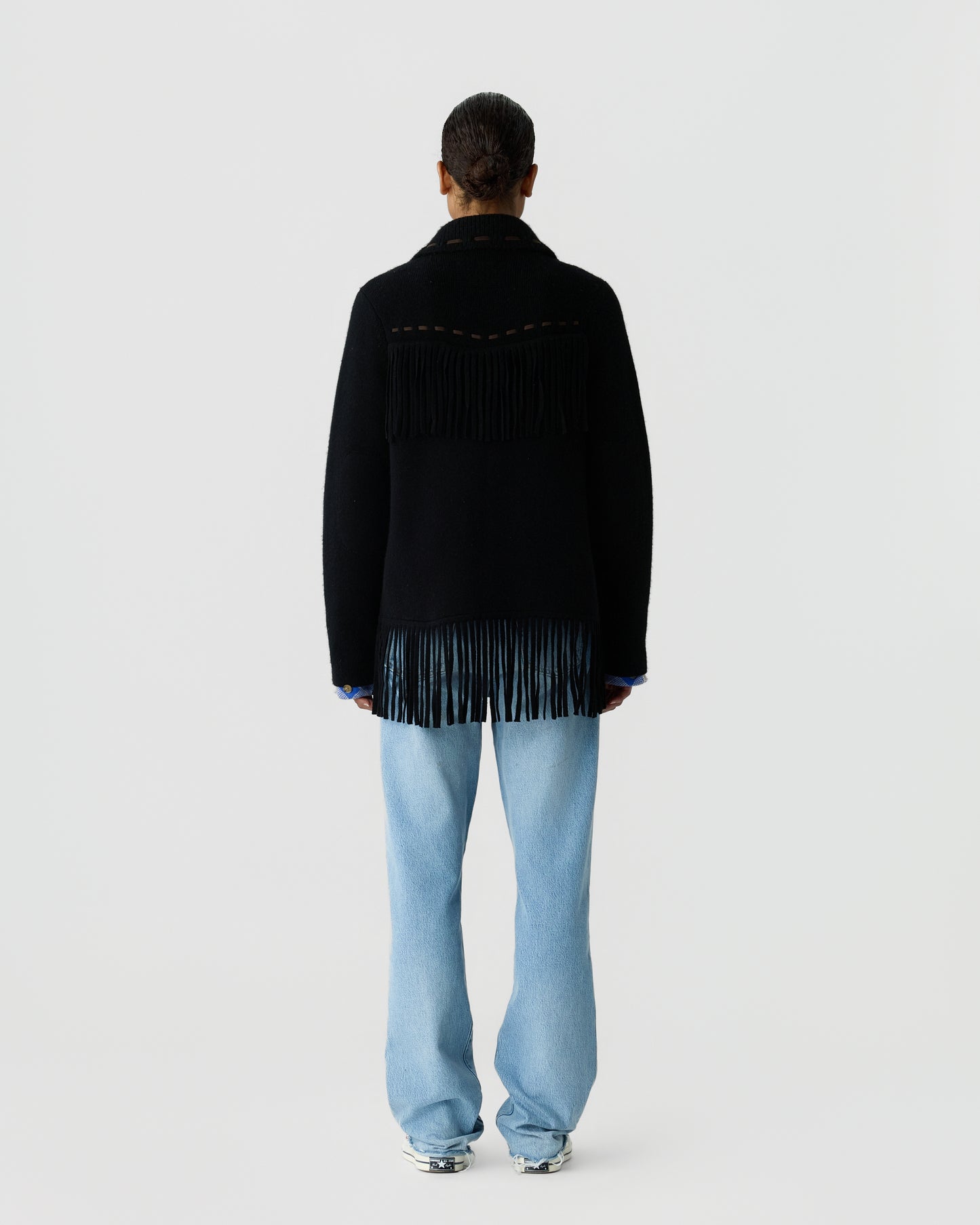 Back view of a person wearing a black fringed sweater with a high collar, light blue wide-leg jeans, and silver shoes against a plain light gray background