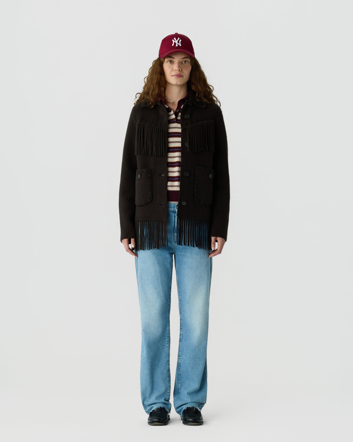 A young woman with curly hair wearing a maroon baseball cap, a black fringed jacket over a striped shirt, light blue straight-leg jeans, and black shoes standing against a plain white background.