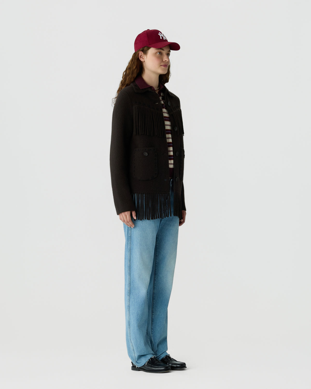 Woman wearing a dark fringed jacket, striped shirt, light blue jeans, black shoes, and a red baseball cap, standing against a plain white background.