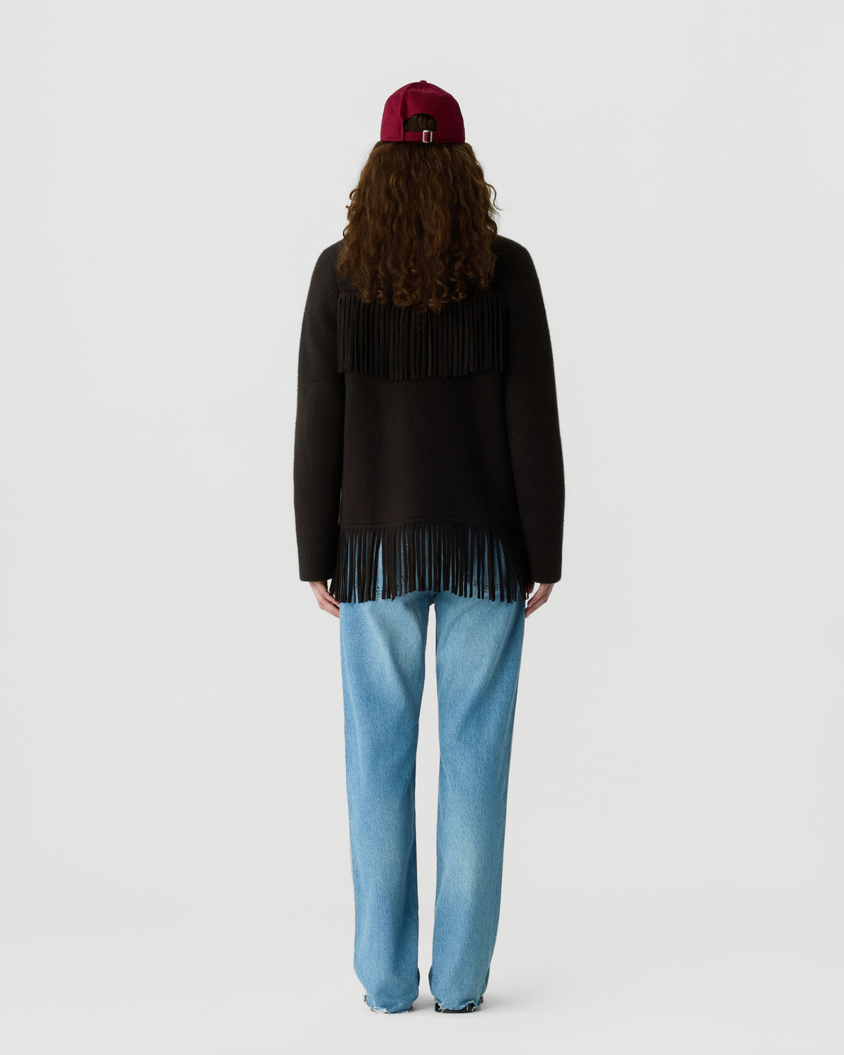 Person with long curly hair wearing a burgundy cap, a dark sweater with fringe details on the back and hem, and light blue jeans standing against a plain background.