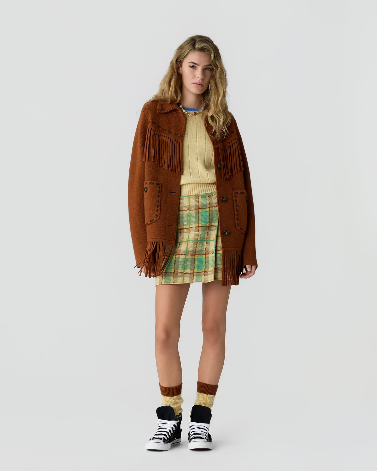 Young woman wearing a brown fringed coat over a yellow sweater and plaid skirt, paired with matching socks and black-and-white sneakers, standing against a plain white background.
