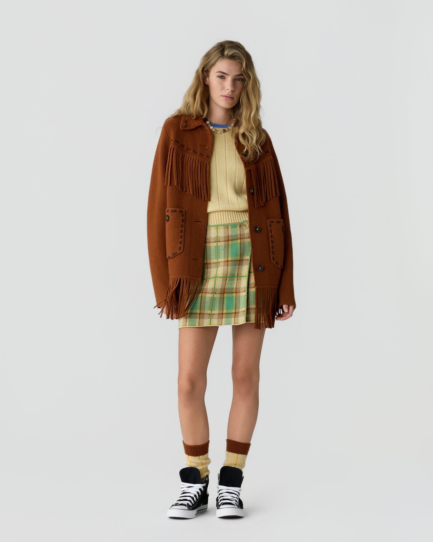 Young woman wearing a brown fringed coat over a yellow sweater and plaid skirt, paired with matching socks and black-and-white sneakers, standing against a plain white background.