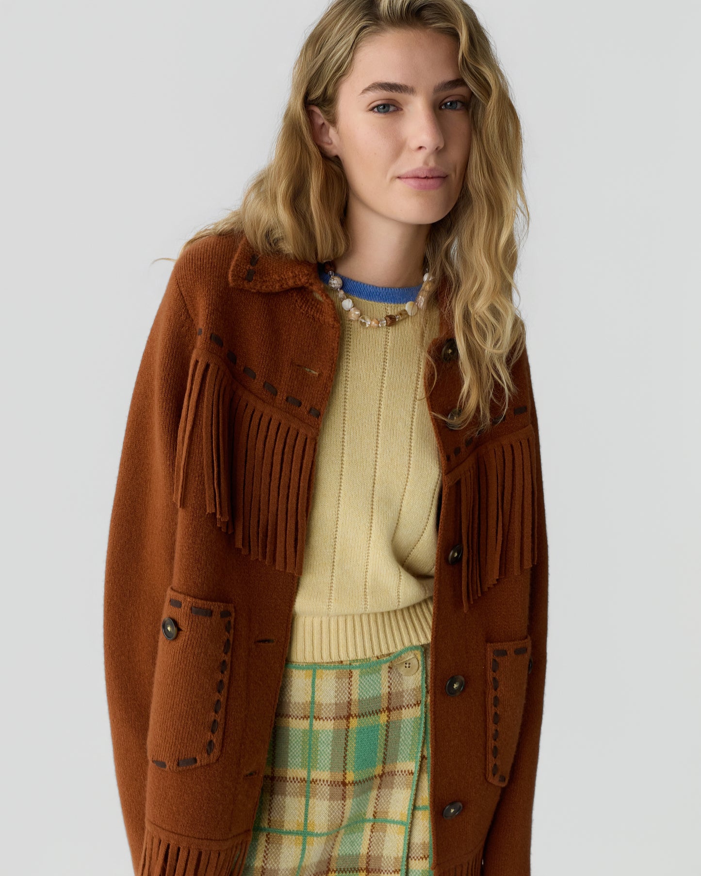 Young woman with wavy blonde hair wearing a brown fringe knit jacket over a beige sweater and green plaid skirt against a light gray background.