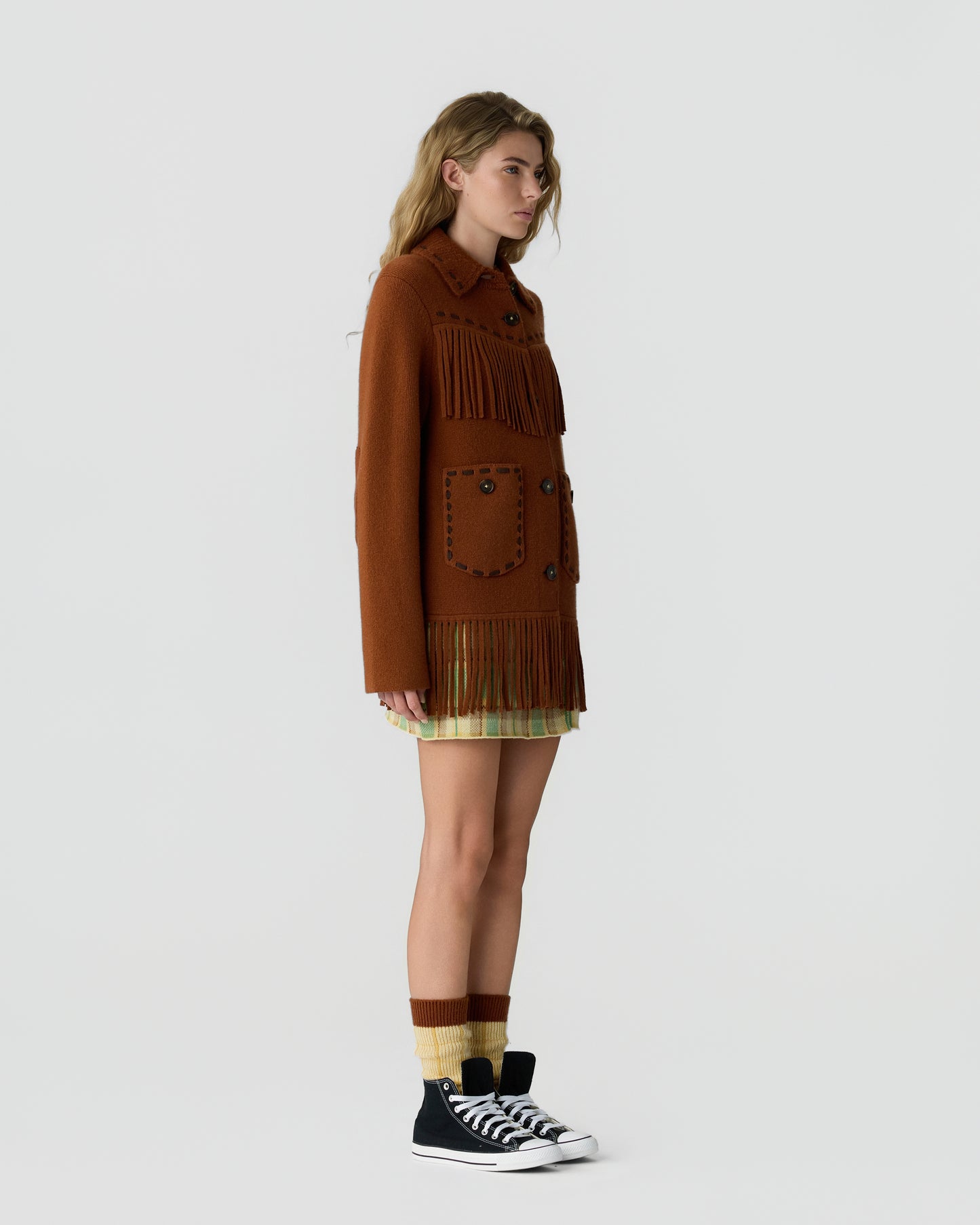 Side view of a woman wearing a brown fringed jacket with button details, a plaid skirt, striped socks, and black high-top sneakers against a plain white background.