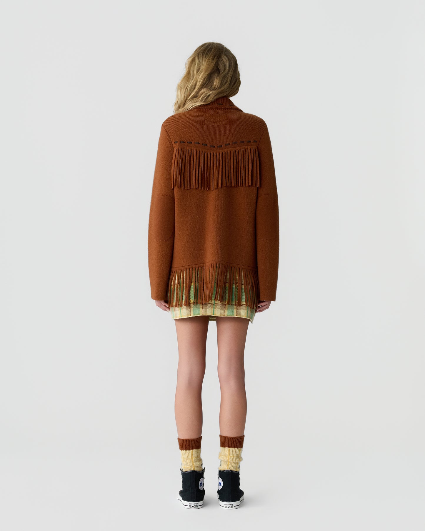 Woman seen from behind wearing a rust-colored fringed sweater over a plaid skirt, paired with mismatched yellow and brown socks and black sneakers on a plain white background.