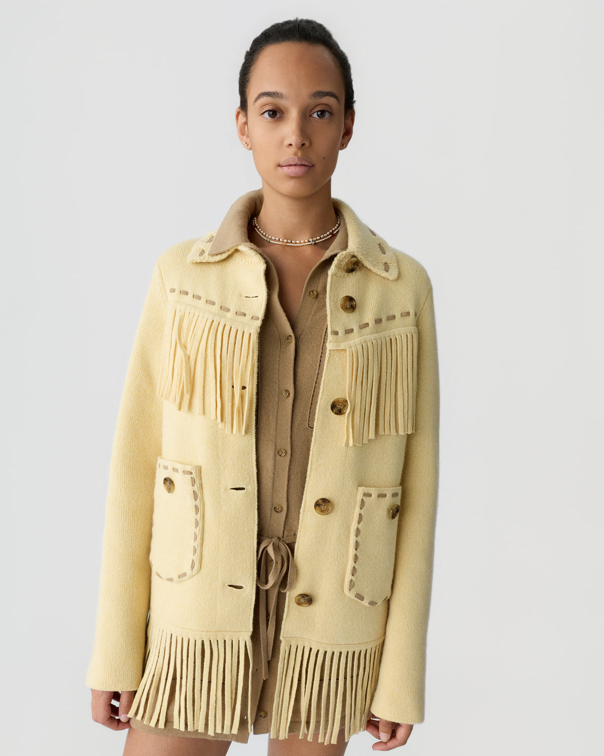 Young woman wearing a beige wool jacket with fringe details and large buttons over a light brown buttoned outfit, standing against a plain gray background.