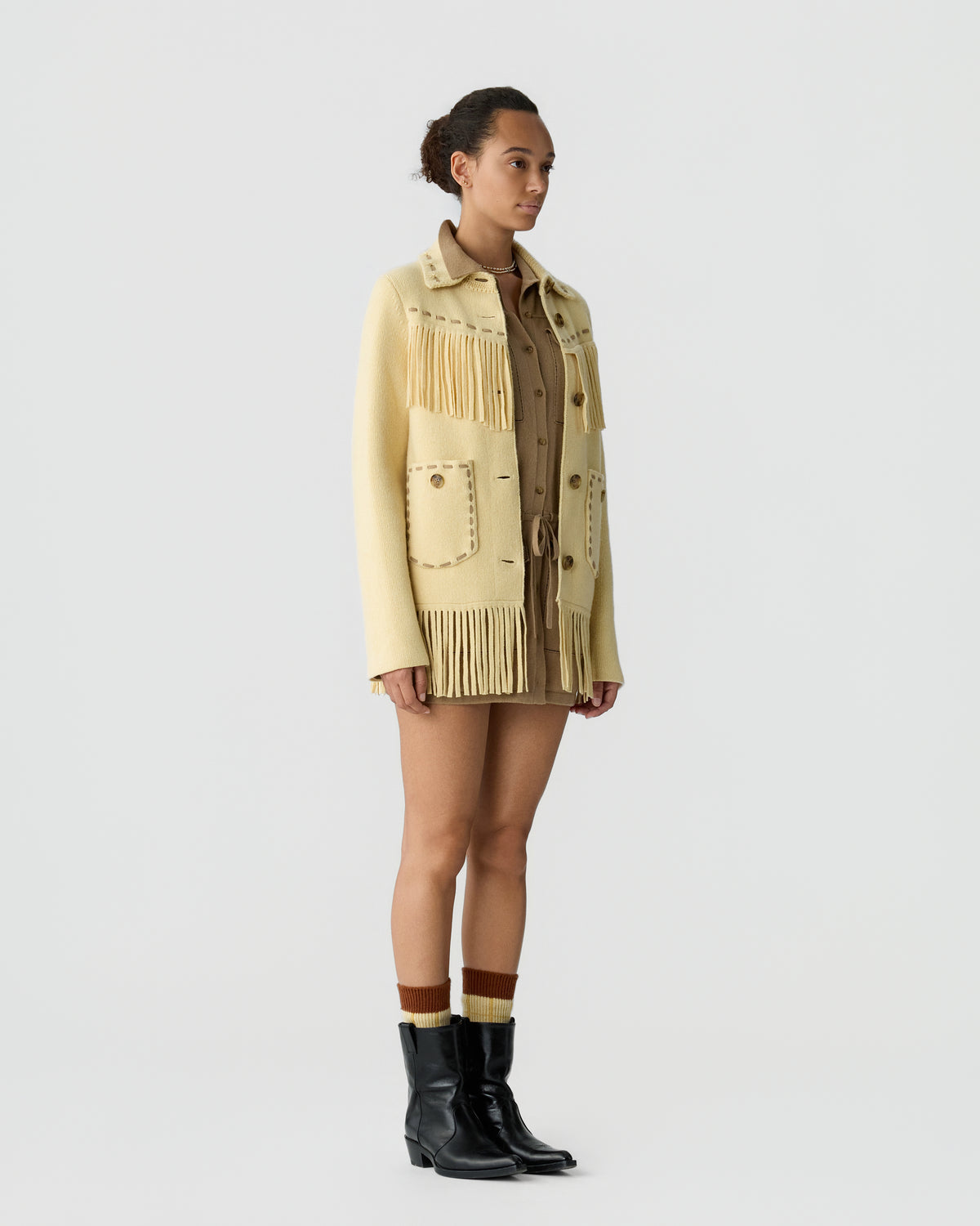 A woman standing against a plain light background wearing a beige fringe jacket over a brown outfit, paired with black ankle boots and striped socks.