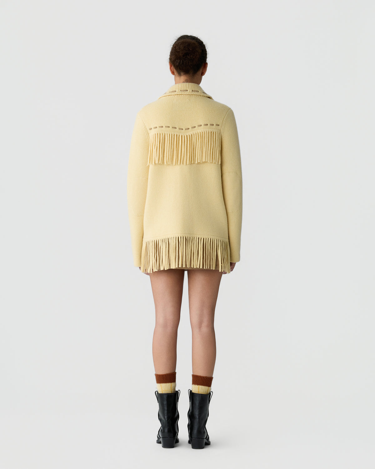 Rear view of a person wearing a light yellow cashmere sweater with long fringe details across the upper back and hem, paired with two-tone socks and black ankle boots.