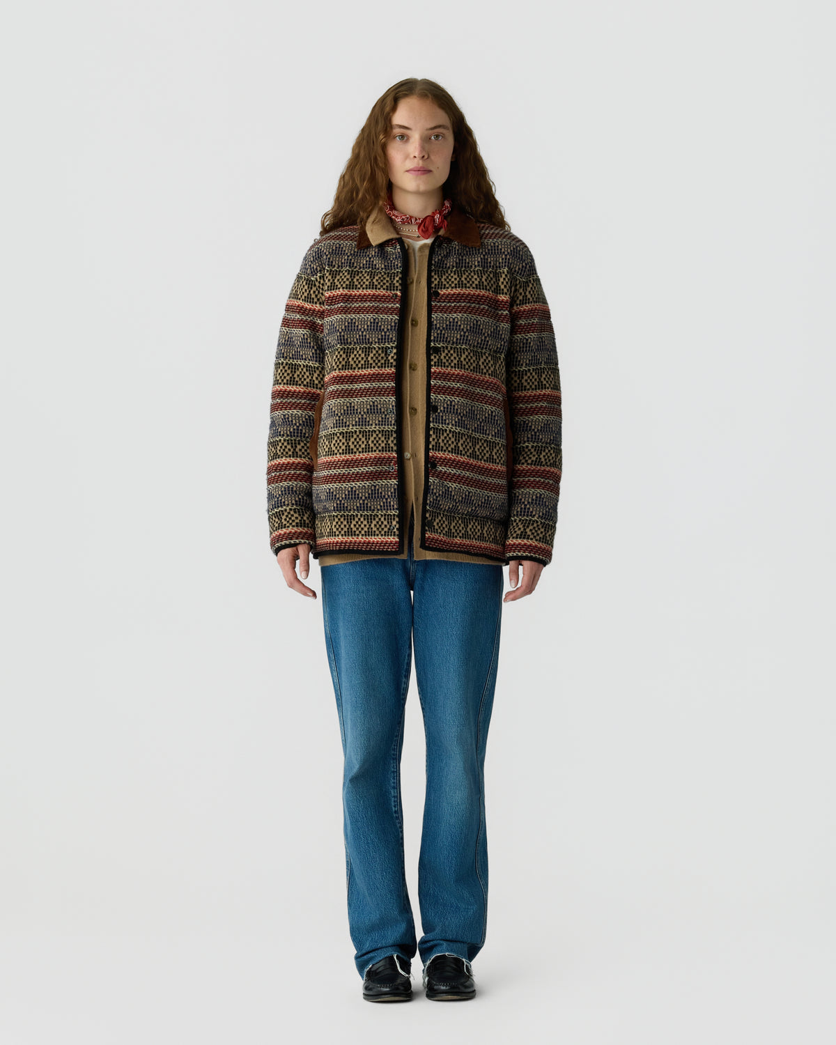 Full length portrait of a woman wearing a patterned zip front cardigan with a red collar, blue jeans, and black shoes against a light gray background