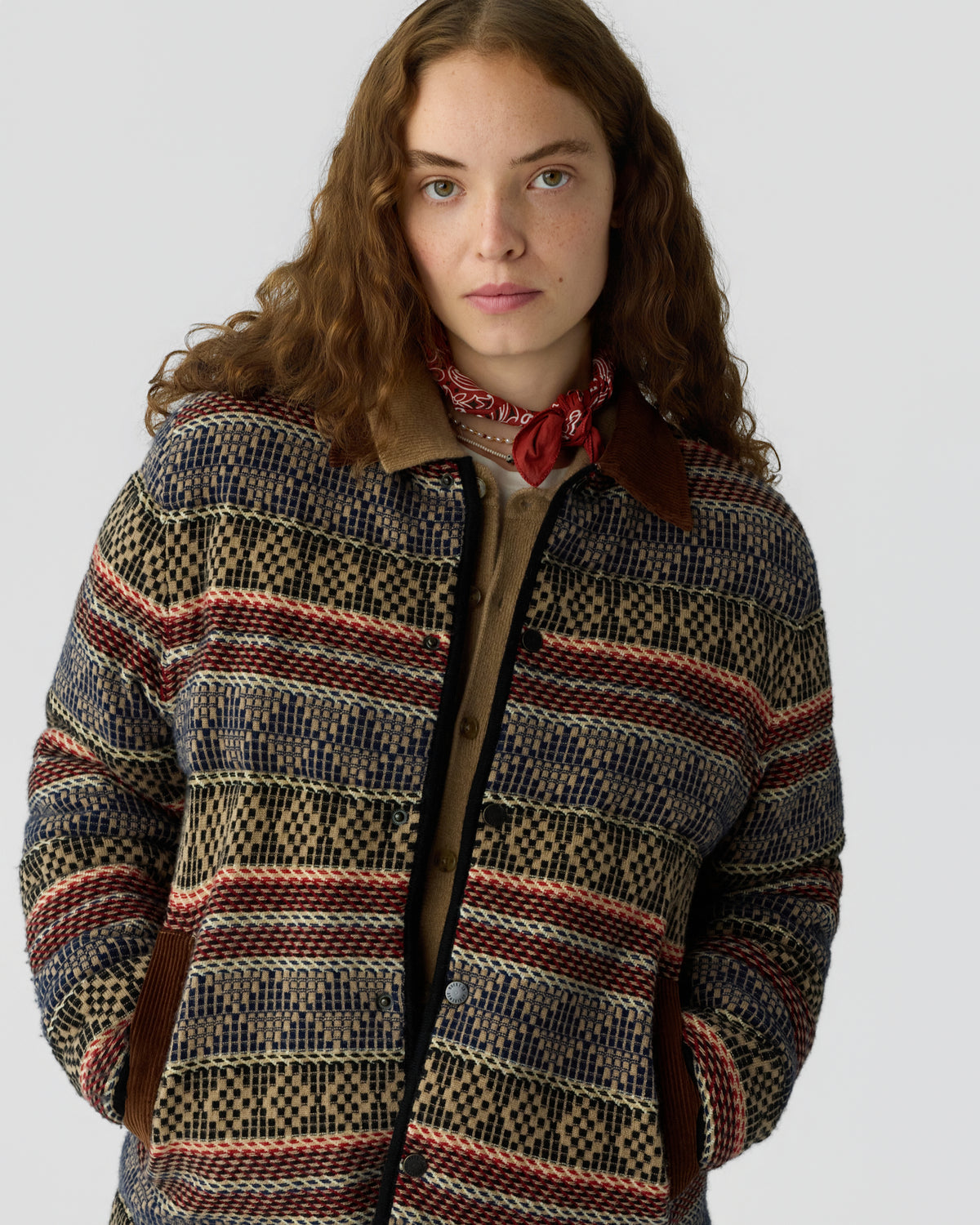 Woman with curly brown hair wearing a patterned button front jacket with horizontal red and beige stripes and a red neck scarf against a light gray background