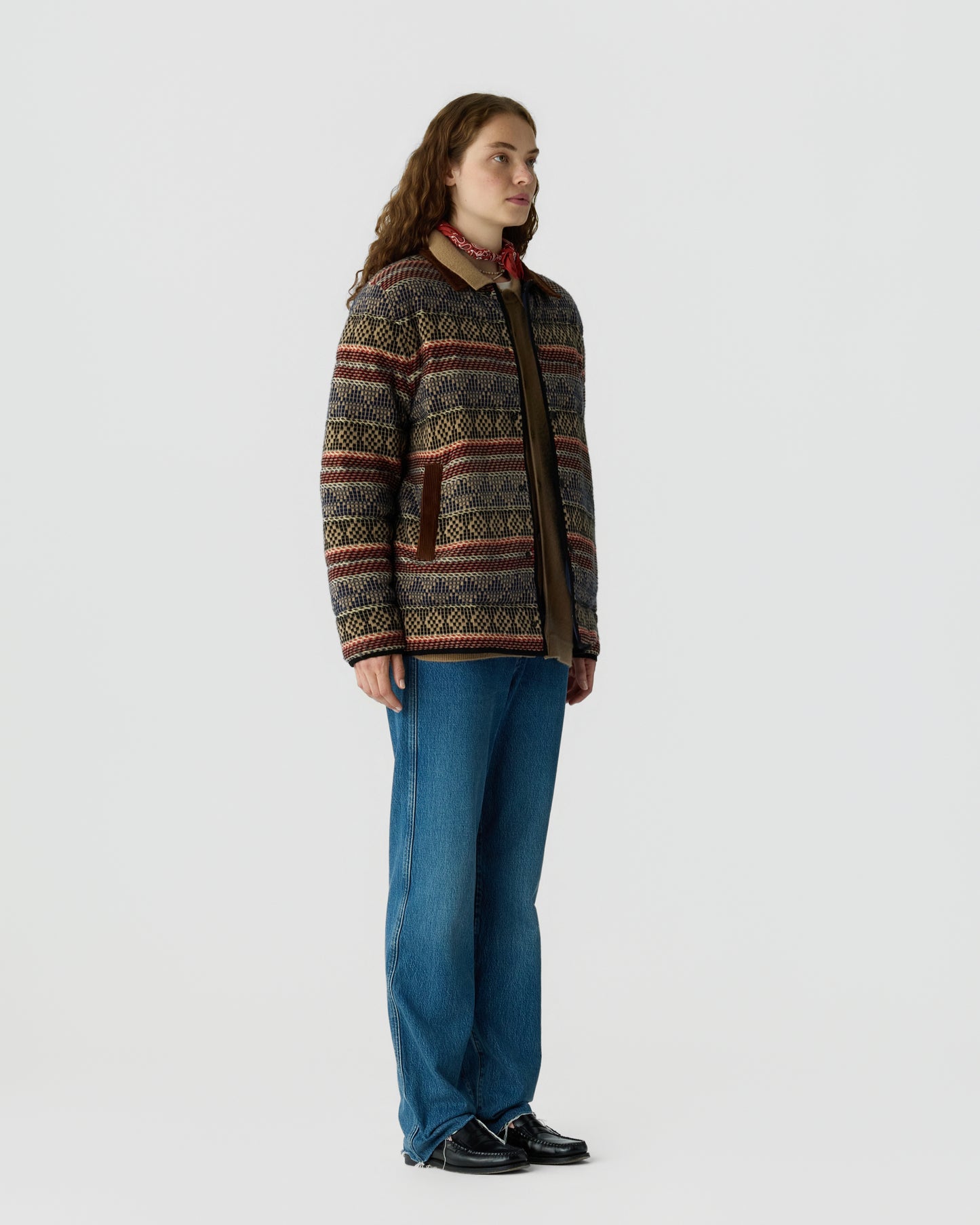 Person wearing a patterned knit cardigan with a red scarf blue wide leg jeans and black shoes against a light gray background