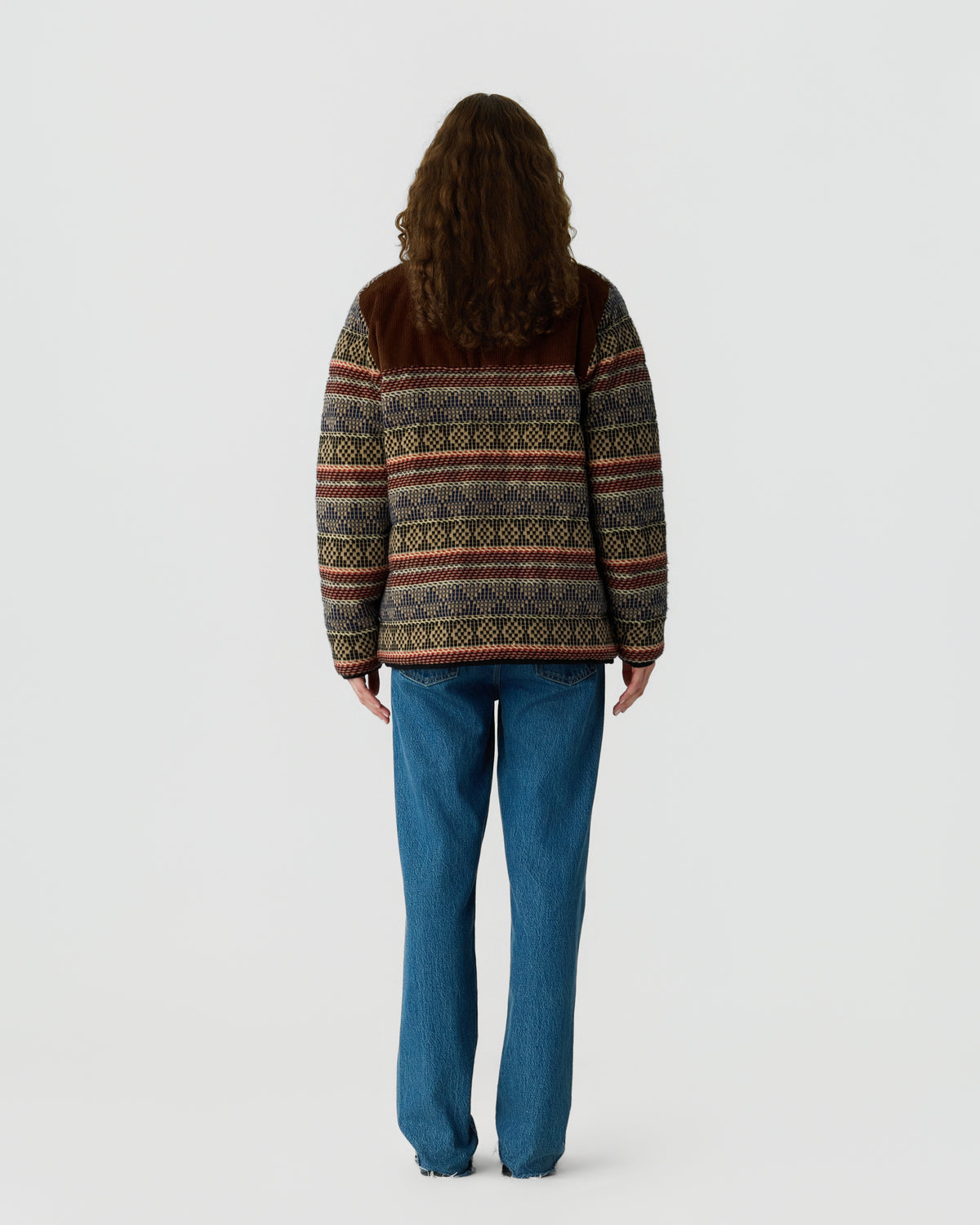 Person viewed from behind wearing a patterned brown and multicolored sweater with blue jeans against a plain light gray background