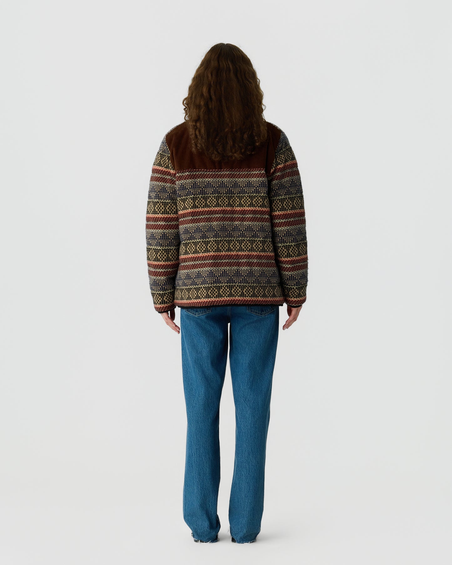 Person viewed from behind wearing a patterned brown and multicolored sweater with blue jeans against a plain light gray background