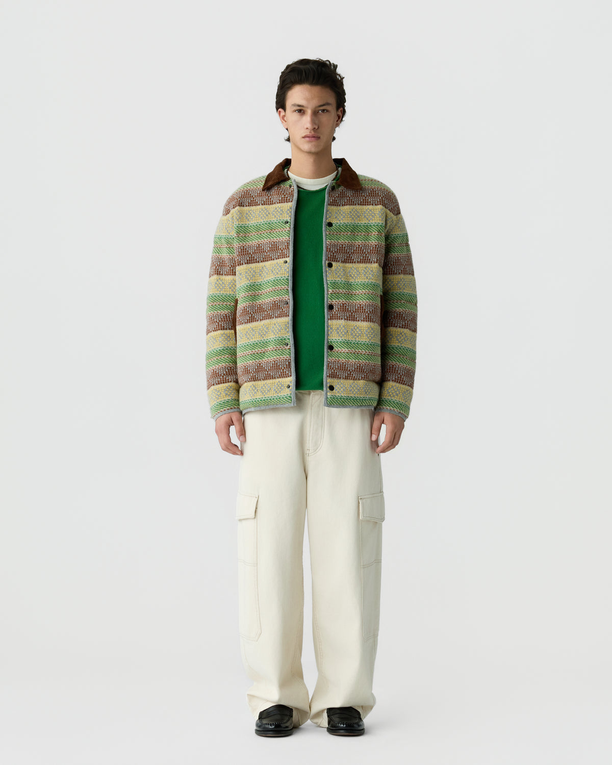 A young man stands against a plain white background wearing a multicolored striped jacket over a green shirt, cream cargo pants, and black shoes.