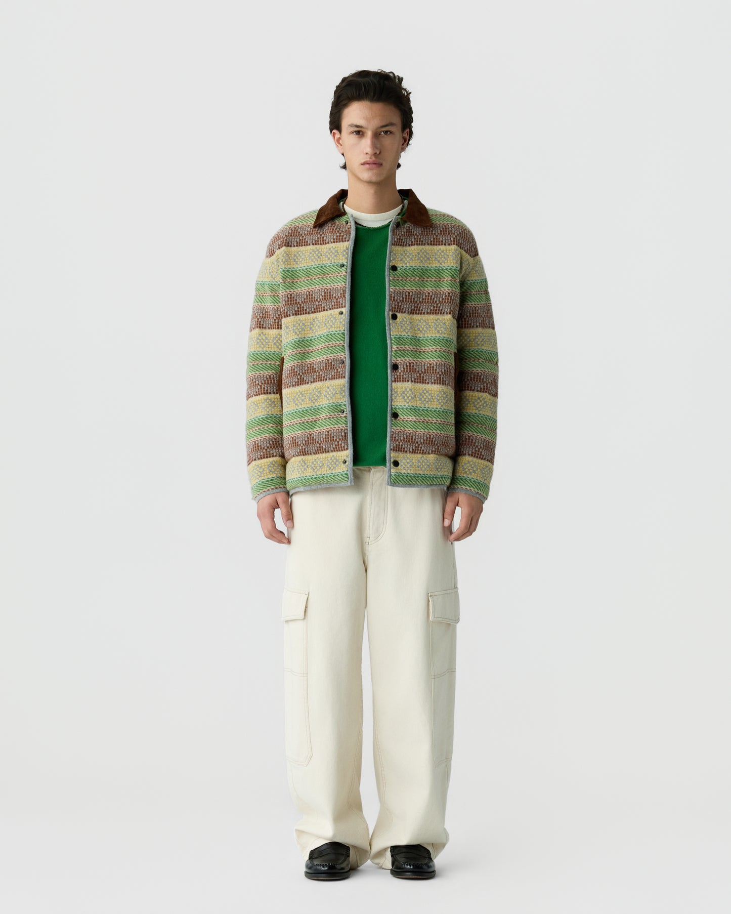 A young man stands against a plain white background wearing a multicolored striped jacket over a green shirt, cream cargo pants, and black shoes.
