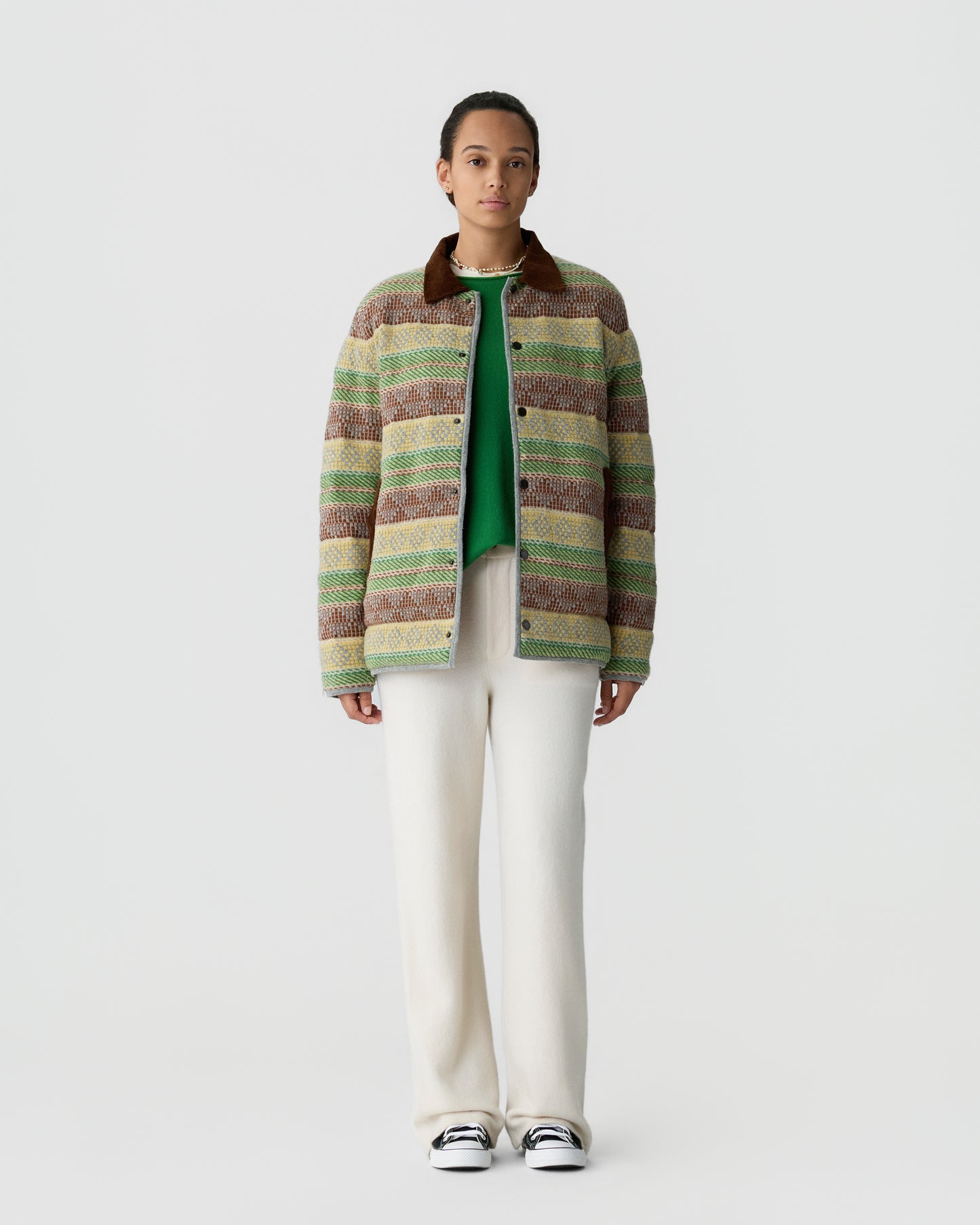 A person stands wearing a multicolored striped knit jacket with a brown collar over a green top, paired with loose white pants and black and white sneakers against a plain white background.