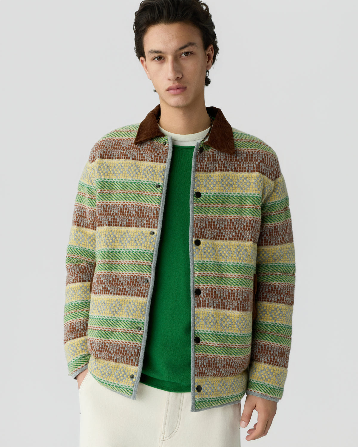 Young man wearing a multicolored striped jacket with a brown collar over a green shirt and light-colored pants, standing against a plain light gray background.